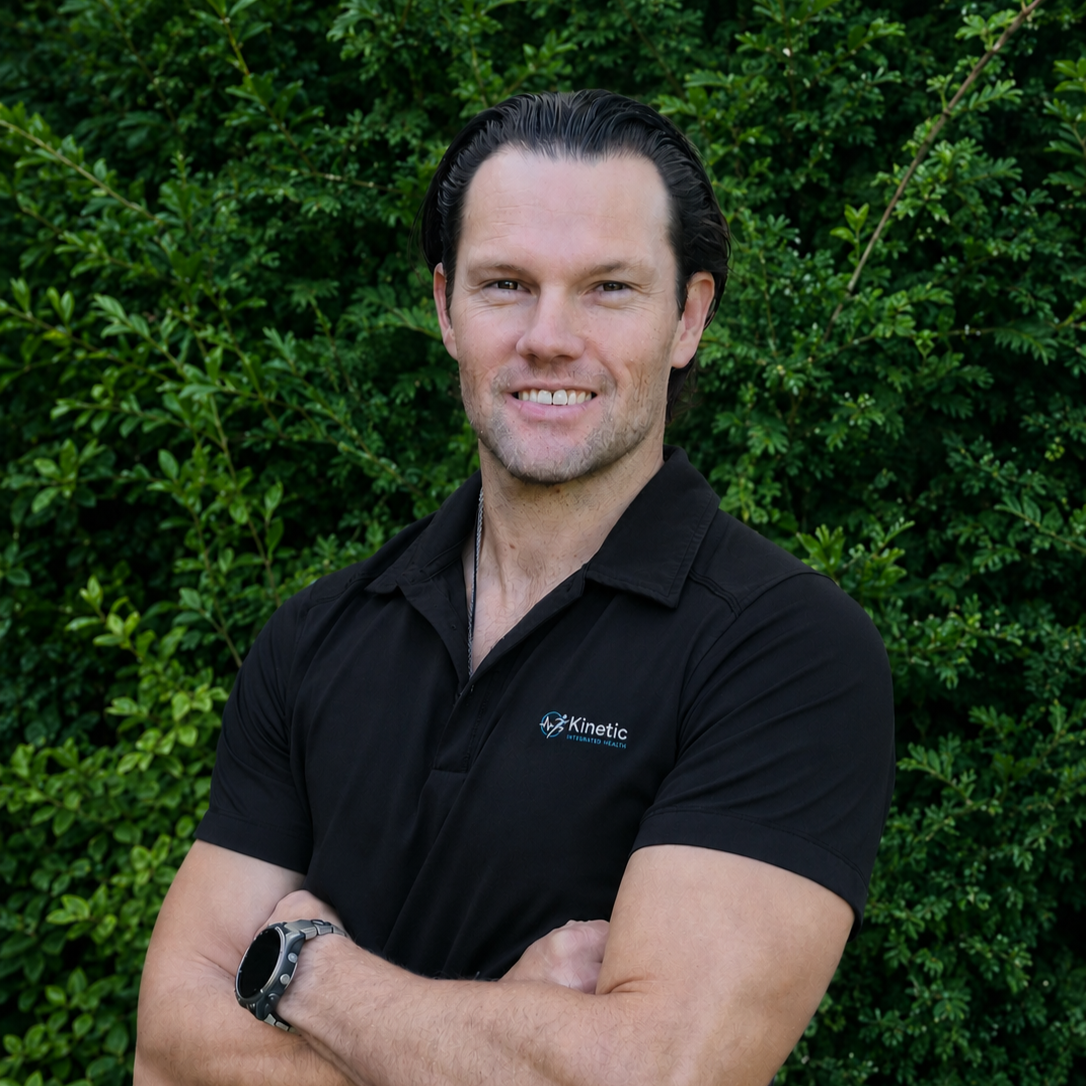 A man standing outdoors with arms crossed, wearing a black polo shirt with a logo, and a wristwatch, smiling in front of green foliage.