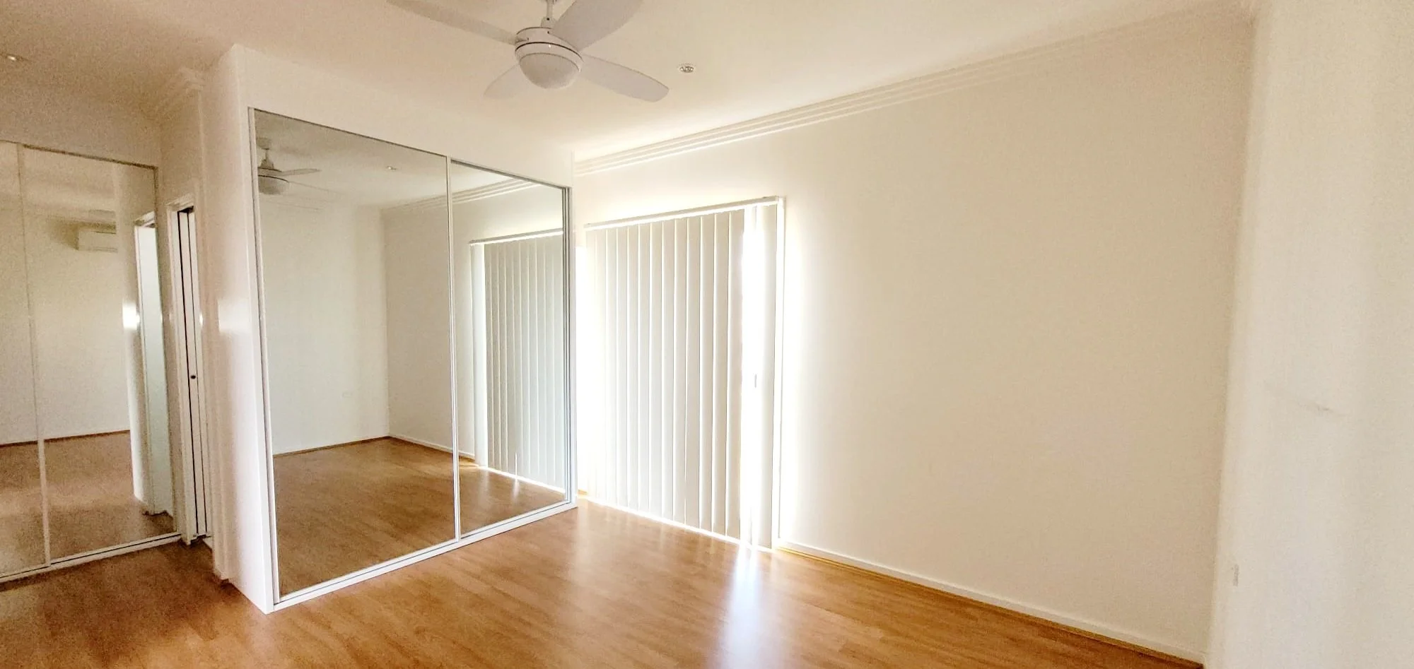 Empty room with wood flooring, white walls, closet with mirrored sliding doors, and vertical blinds covering a window or door, lit with natural light.