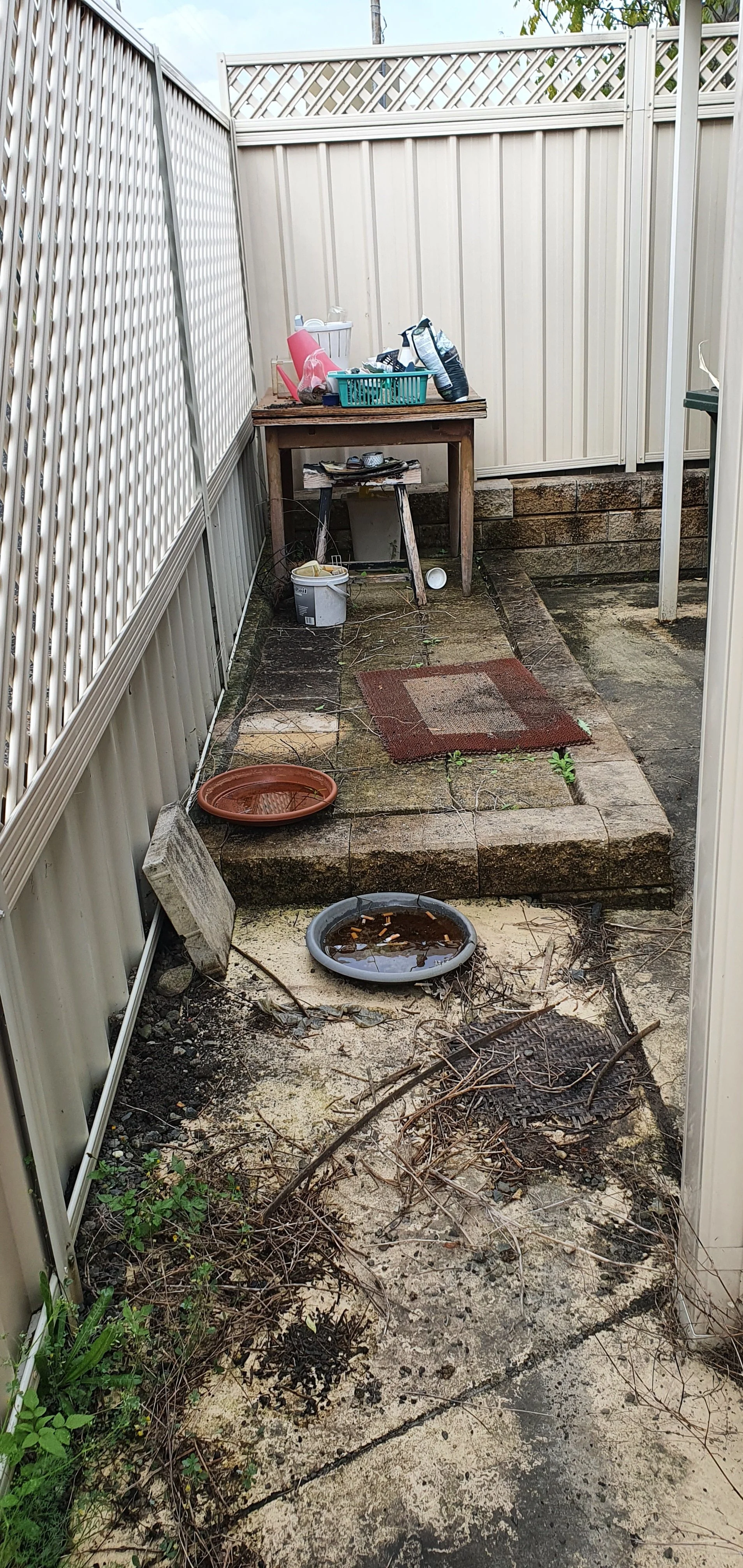 A small, cluttered outdoor patio area with an uneven surface. There are two shallow bowls filled with water or debris on the ground, among scattered twigs and weeds. A raised brick platform is present, with a small wooden table on top filled with var