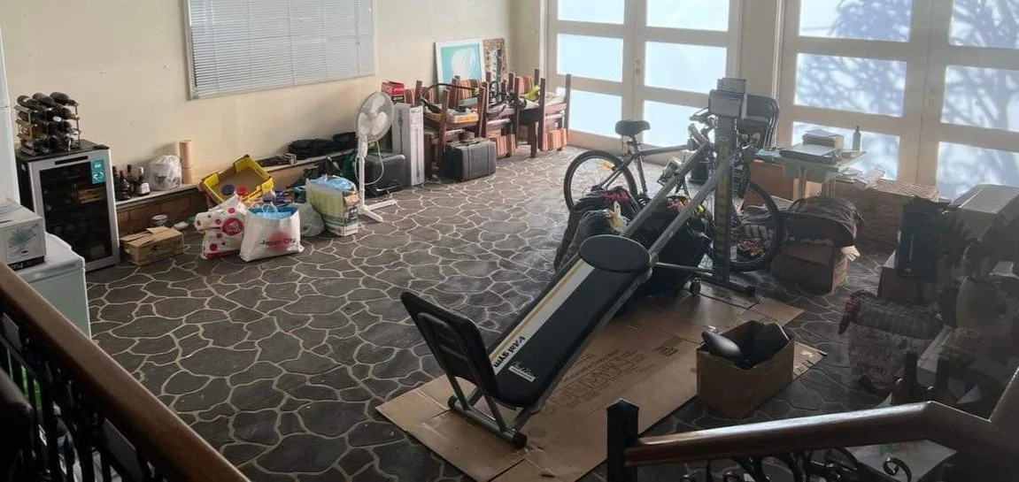 Room with various household items, furniture, and exercise equipment including a treadmill, bicycles, and chairs, with sunlight coming through large windows.