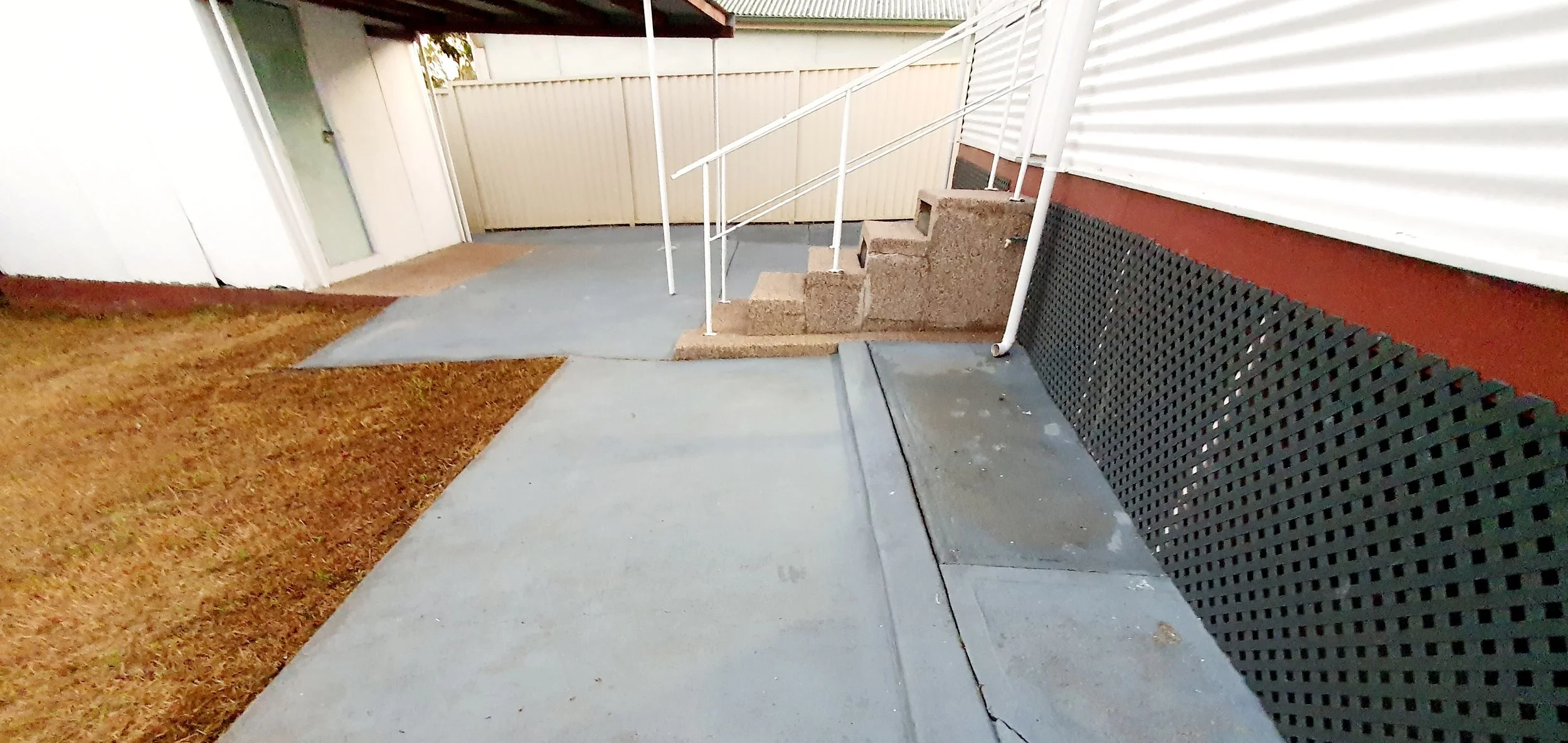 Concrete walkway with gravel stairs leading to the house entry. White railing on stairs. Brown lattice skirt on house wall. Brown and white siding. Fence in background.