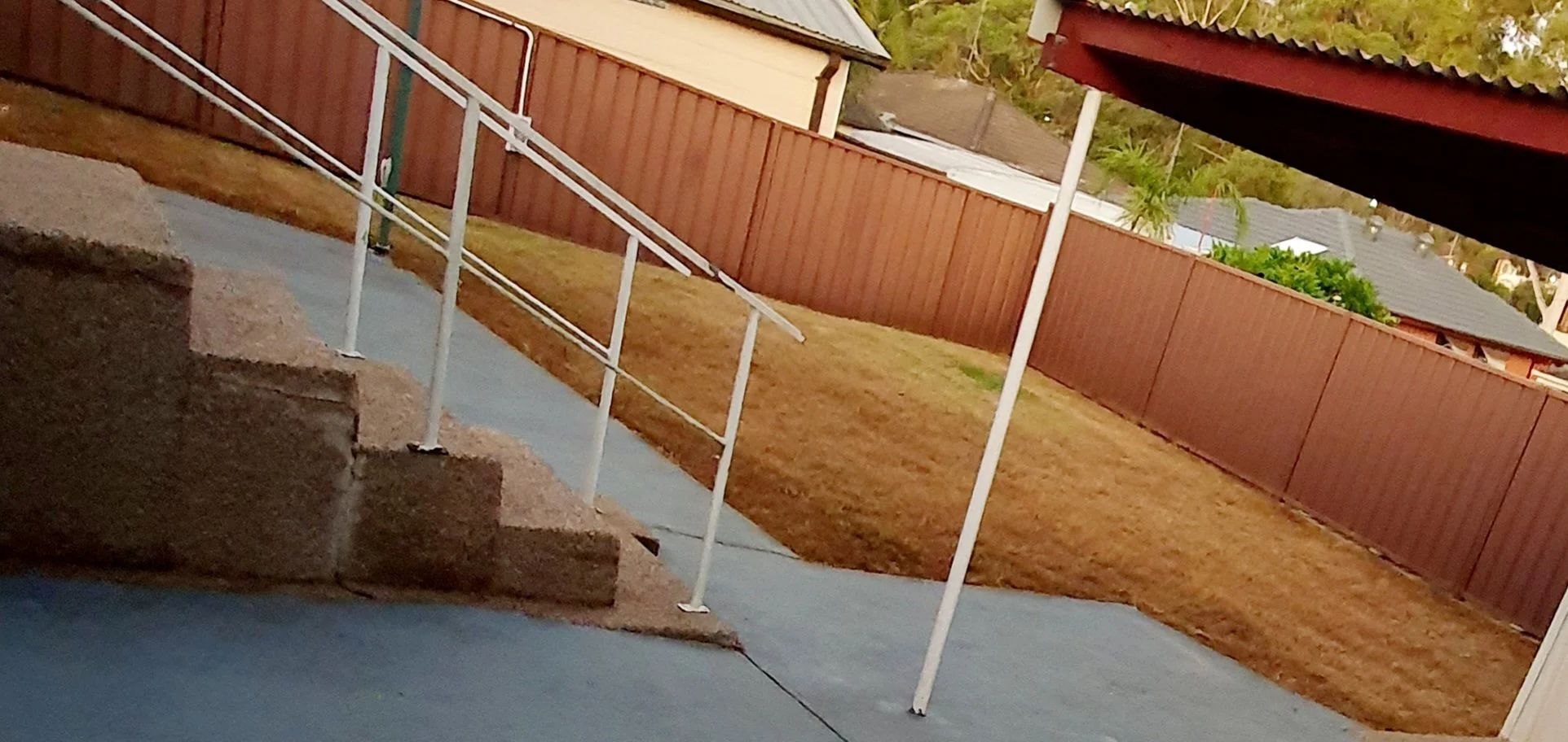 Backyard area with stairs and white handrails, brown fencing, and a cemented patio with a grassy yard.