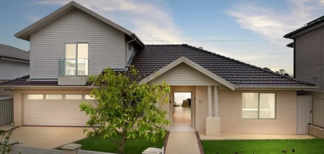A two-story house with a gabled roof, brick and vinyl siding exterior, a two-car garage, a front door flanked by columns, and a front yard with a small tree and grass.