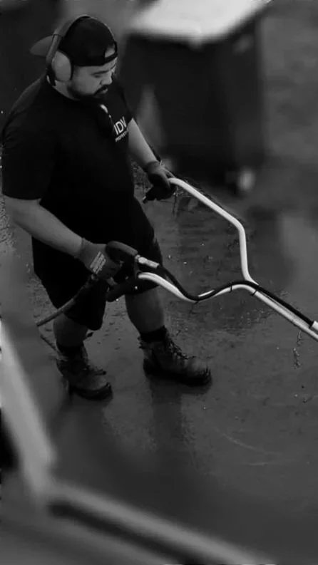 A man wearing headphones, a cap, and a black shirt with white text working outdoors with a power tool on a paved surface.