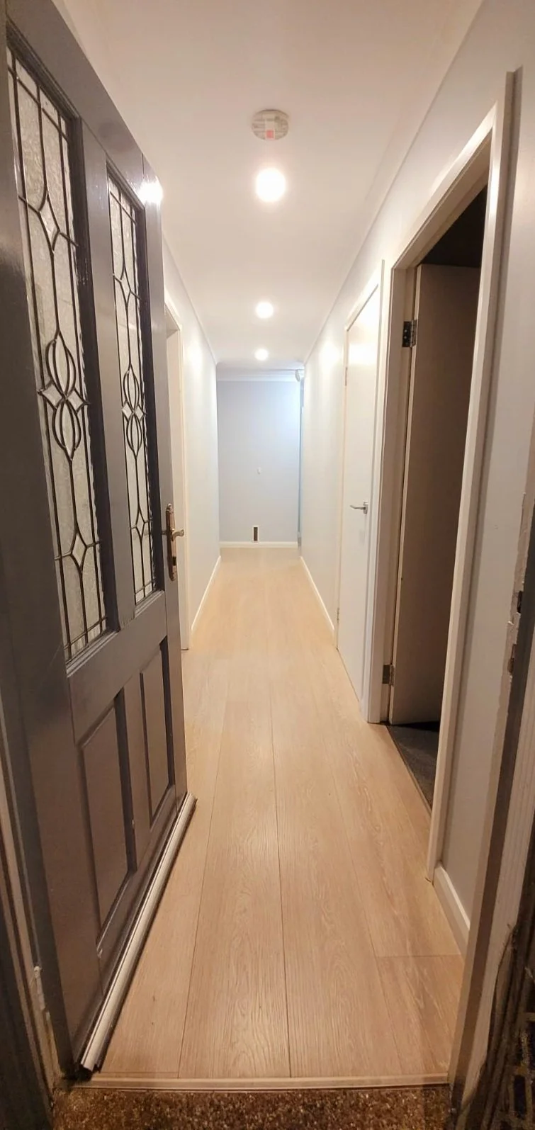 View of a narrow hallway with wooden flooring, white walls, ceiling lights, and multiple closed white doors, with a section of a decorative glass door on the left.