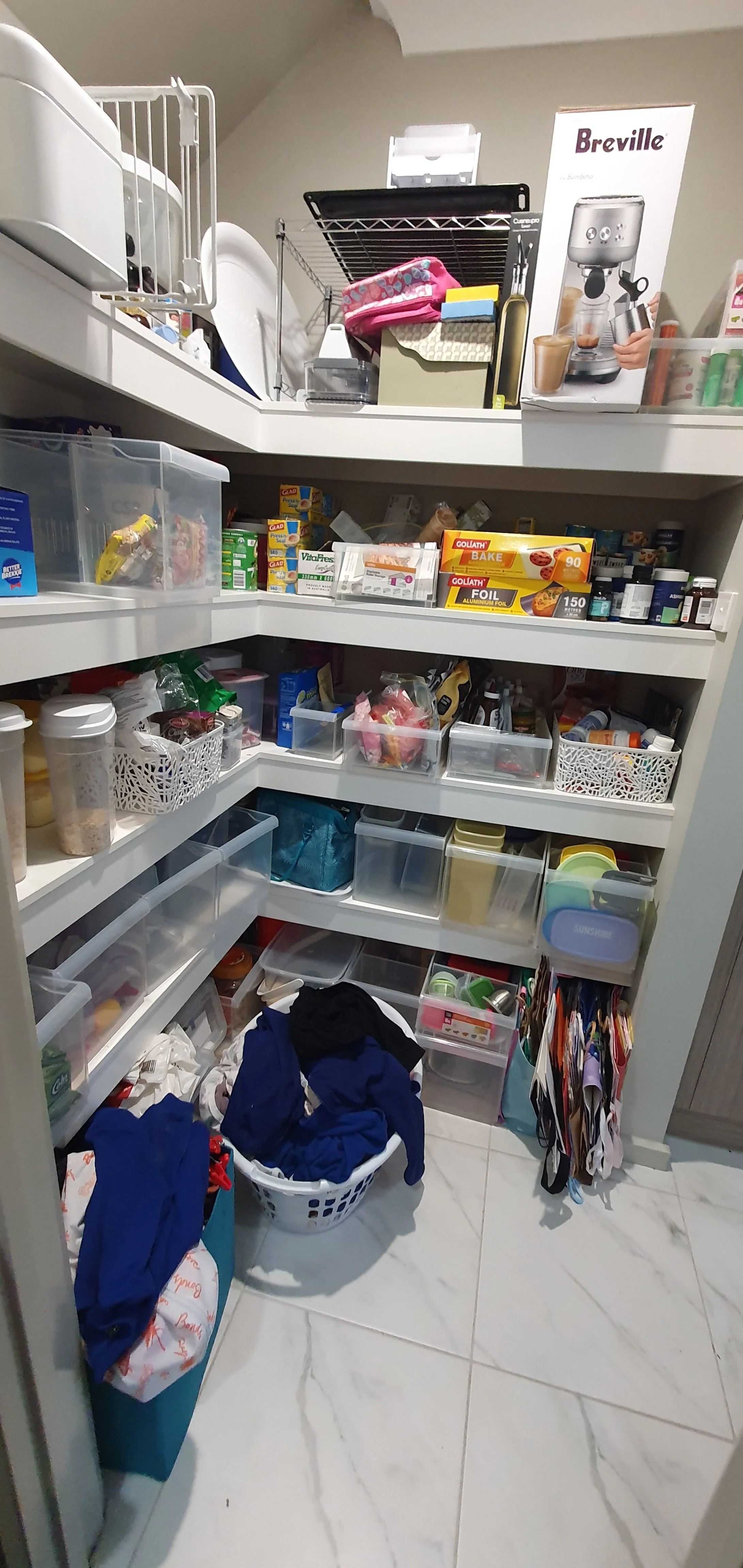 Cluttered pantry with various food items, containers, and miscellaneous household items on white shelves, with laundry basket and grocery bag on the tiled floor.