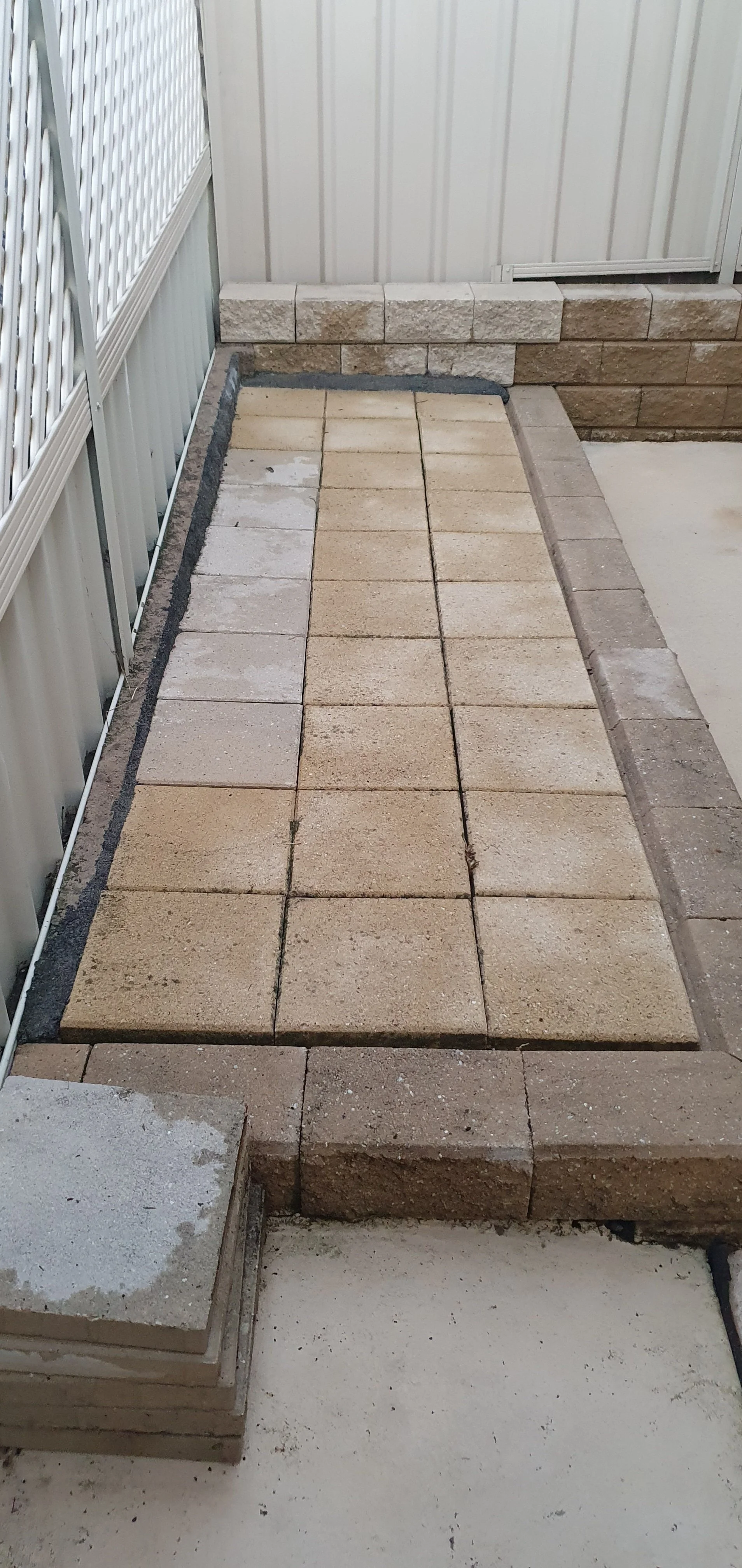 A small outdoor area with a partially completed brick paver walkway, bordered by a white metal fence and a beige metal shed wall.