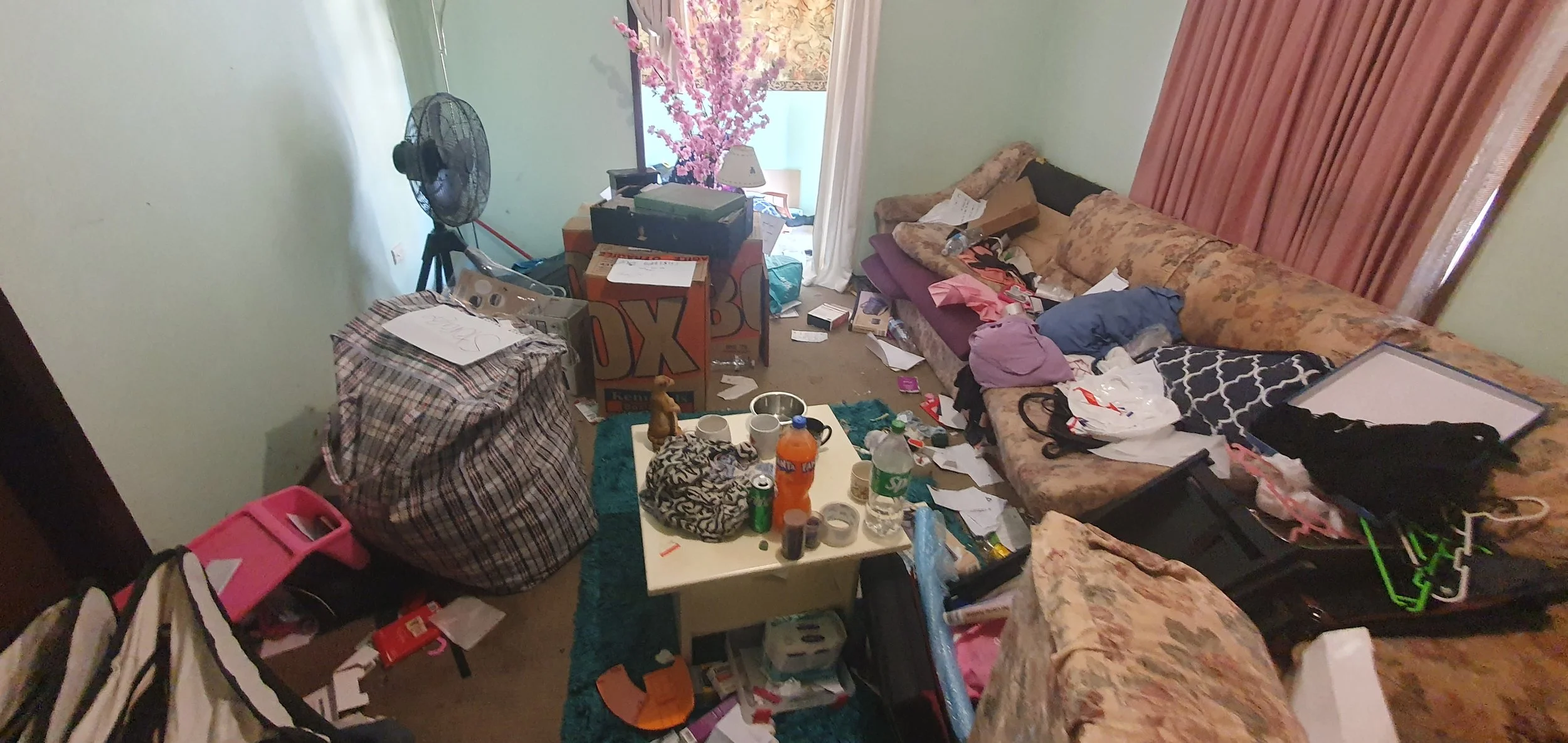 Cluttered living room with messy couches, scattered papers, bottles, and boxes, a fan in the corner, and a small table with items.