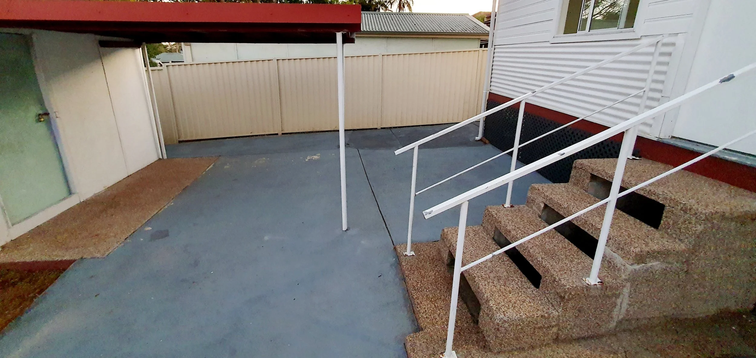 Back patio area with concrete floor, stairs with white metal railing leading to an elevated deck, beige security gate, and part of a house with a green door and a window.