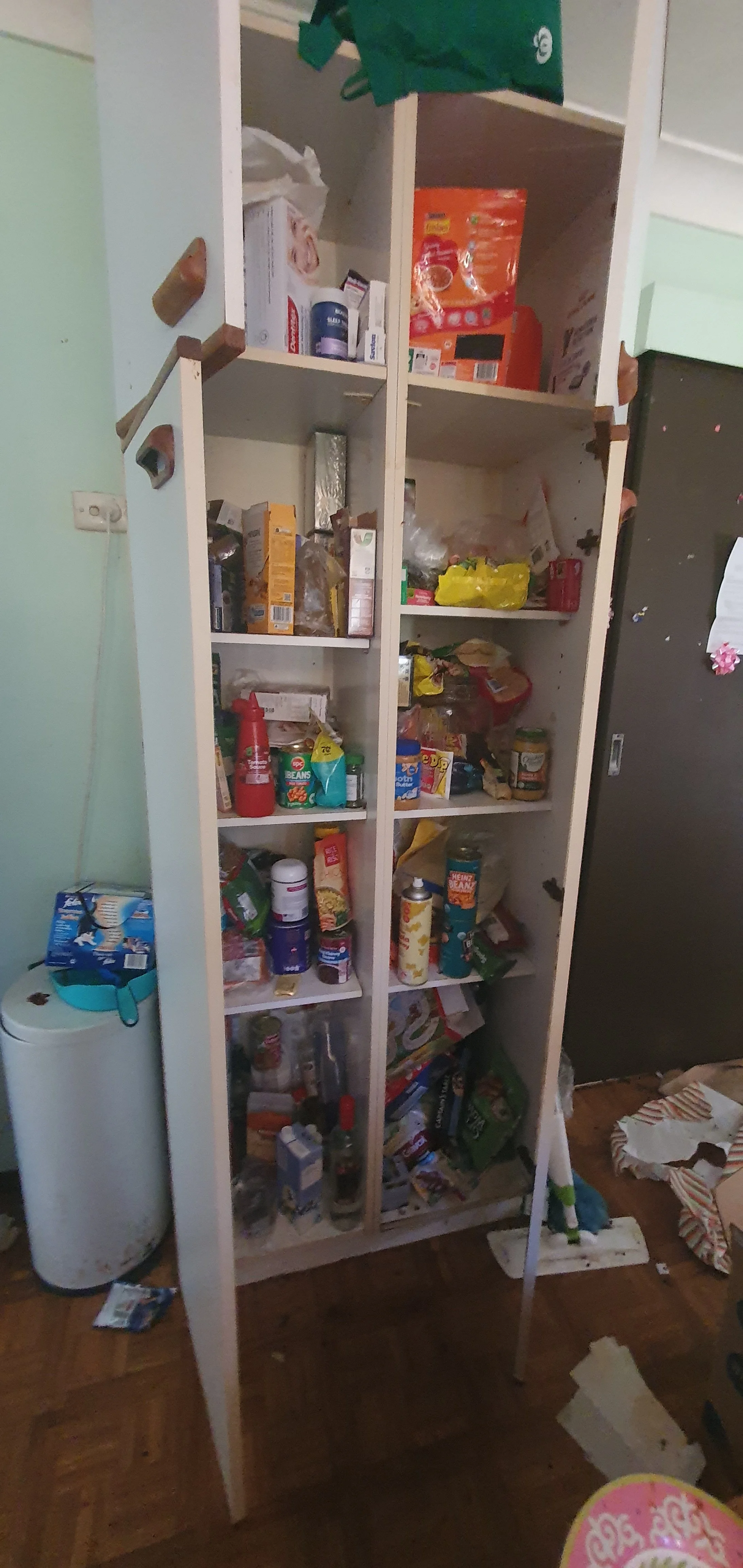 A white kitchen pantry with open shelves filled with snacks, canned goods, and various food items. The pantry door is missing, and there are clutter and boxes around it on a wooden floor.