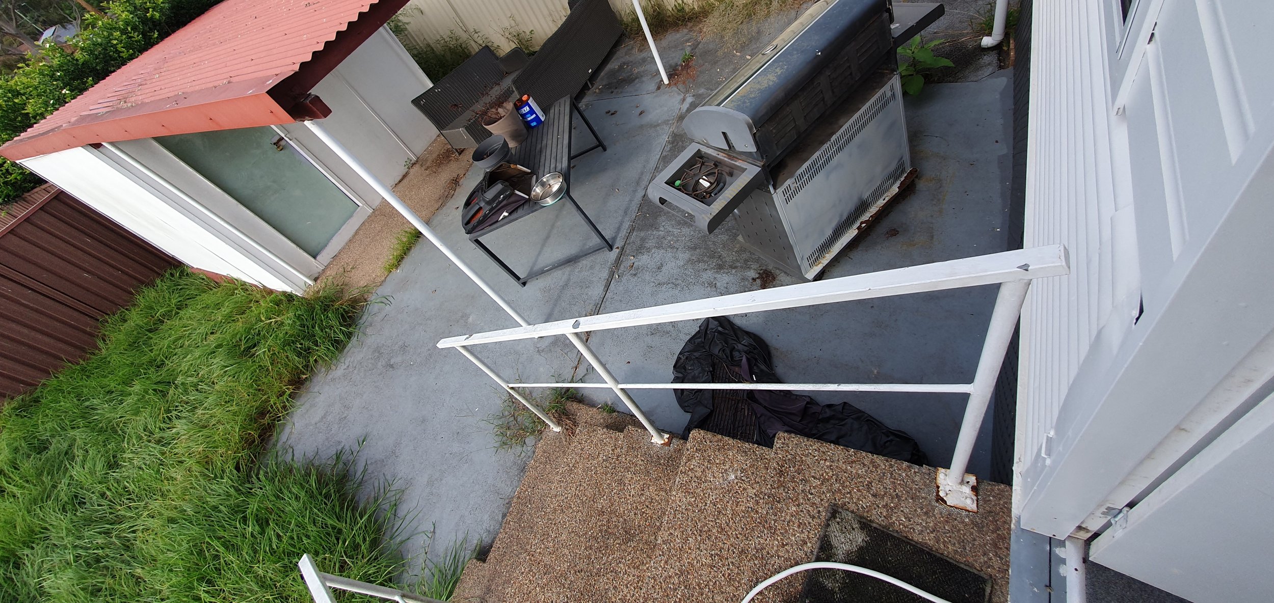 Backyard patio with a small table, chairs, barbecue grill, and a black cover on the ground, next to a white house with a screen door, green plants, and a grassy area.