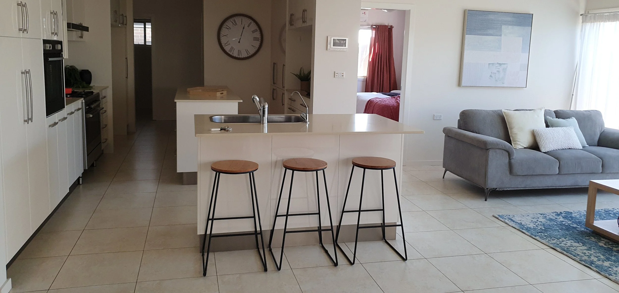 Open-plan living room and kitchen with beige tiles, white cabinetry, a kitchen island with a sink and three barstools, a gray sofa with pillows, and a wall clock.