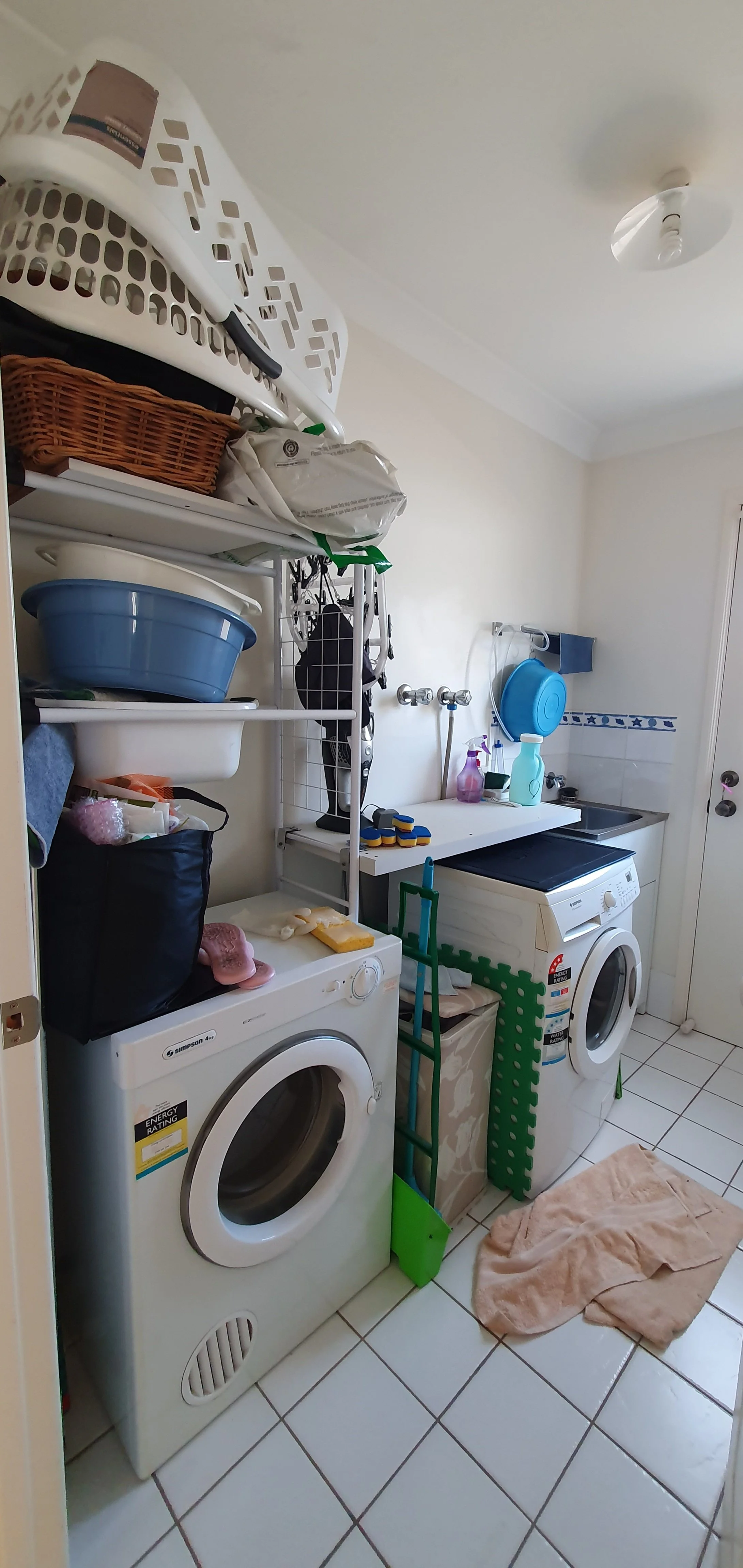 A small utility room with laundry appliances, shelving, and cleaning supplies. There are two washing machines, a sink, and various baskets and items on the shelves.