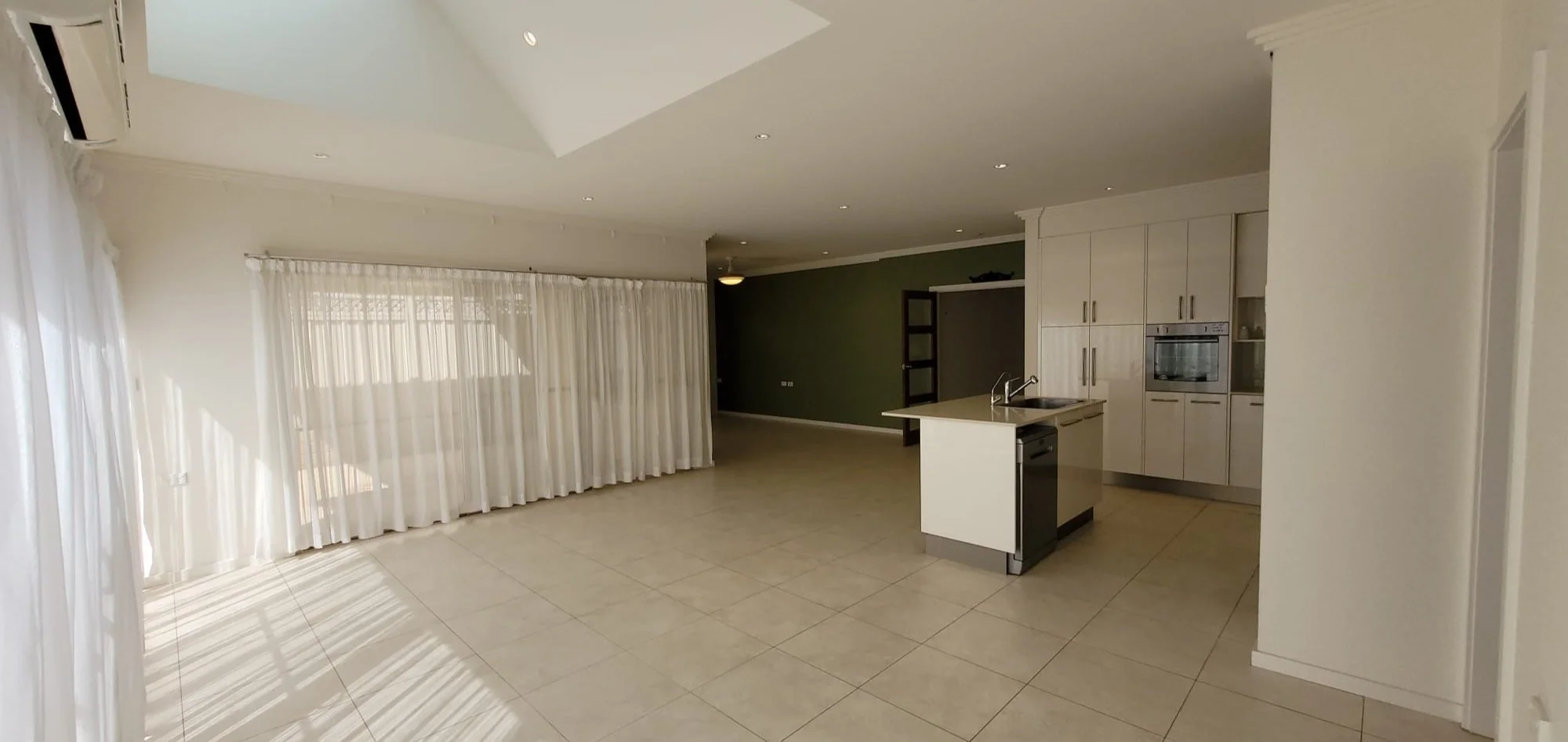 Empty living room and kitchen area with white cabinets, a small island with a sink, large windows with white curtains, and tiled floors.