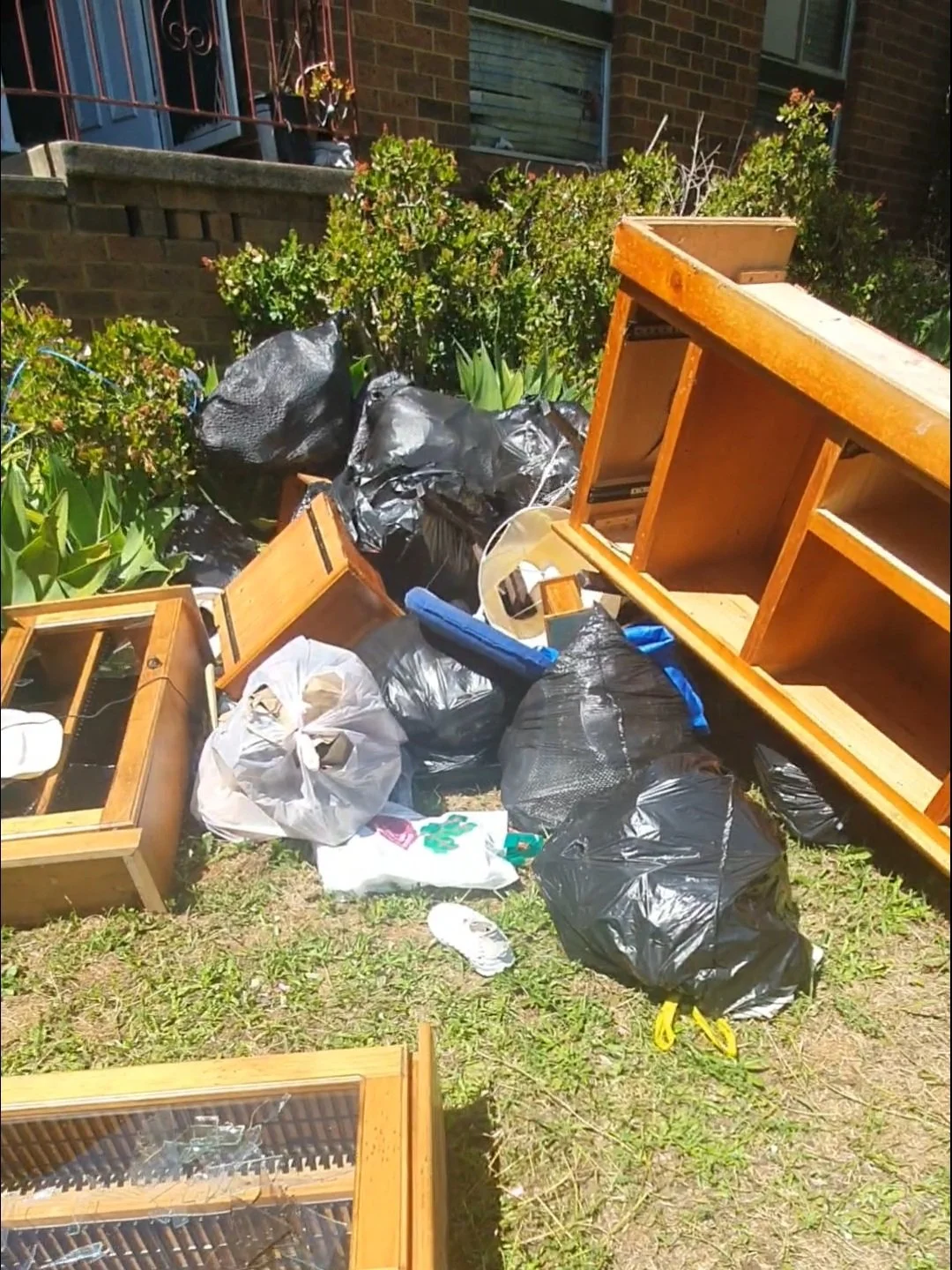 Disorganized pile of trash and discarded furniture on a patch of grass outside a house, including black garbage bags, broken wooden furniture, and a pair of white sneakers.