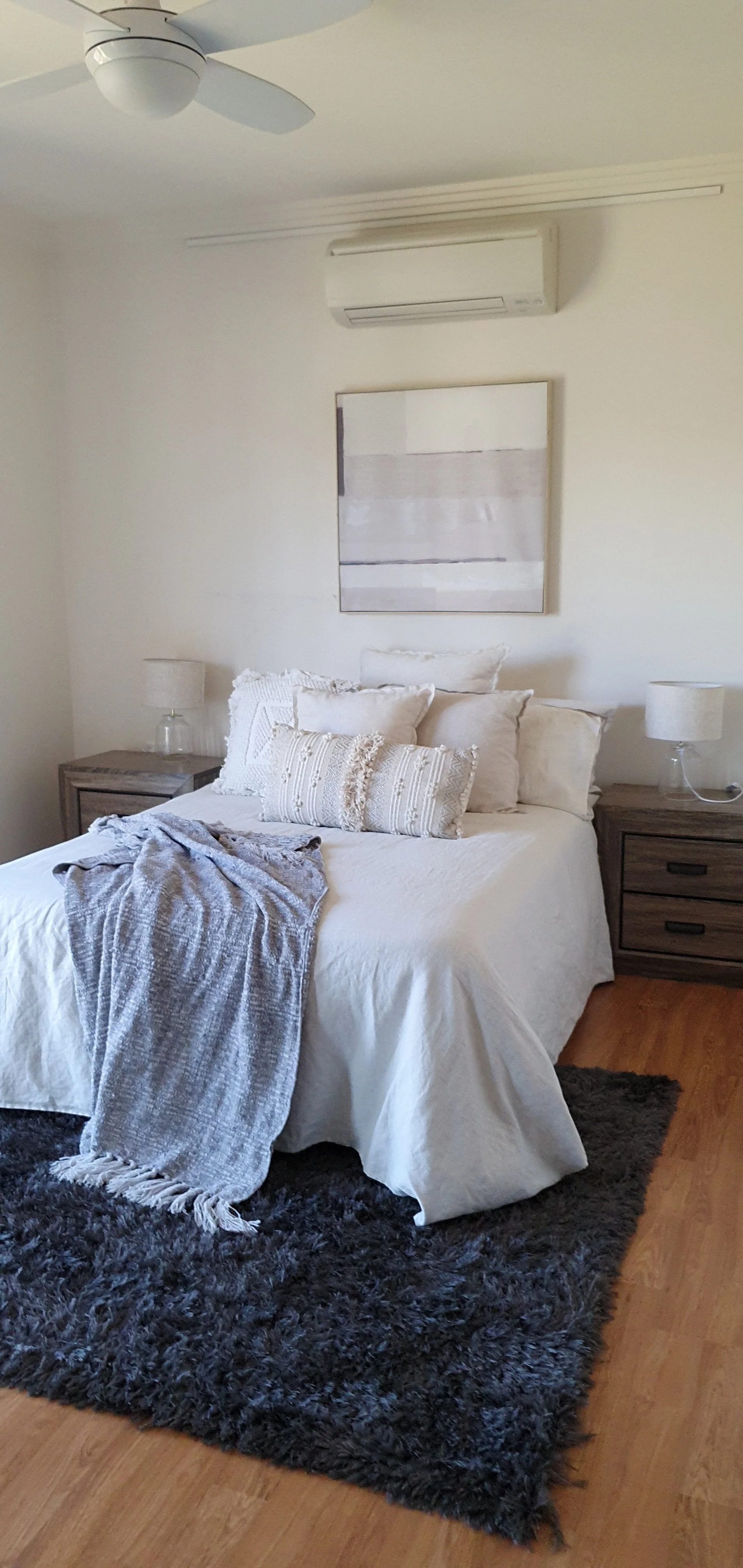 A bedroom with a white bed, neutral-colored pillows, a gray throw blanket, wooden nightstands with lamps, a abstract wall art above the bed, a ceiling fan, an air conditioning unit, and a black shag rug on a wooden floor.