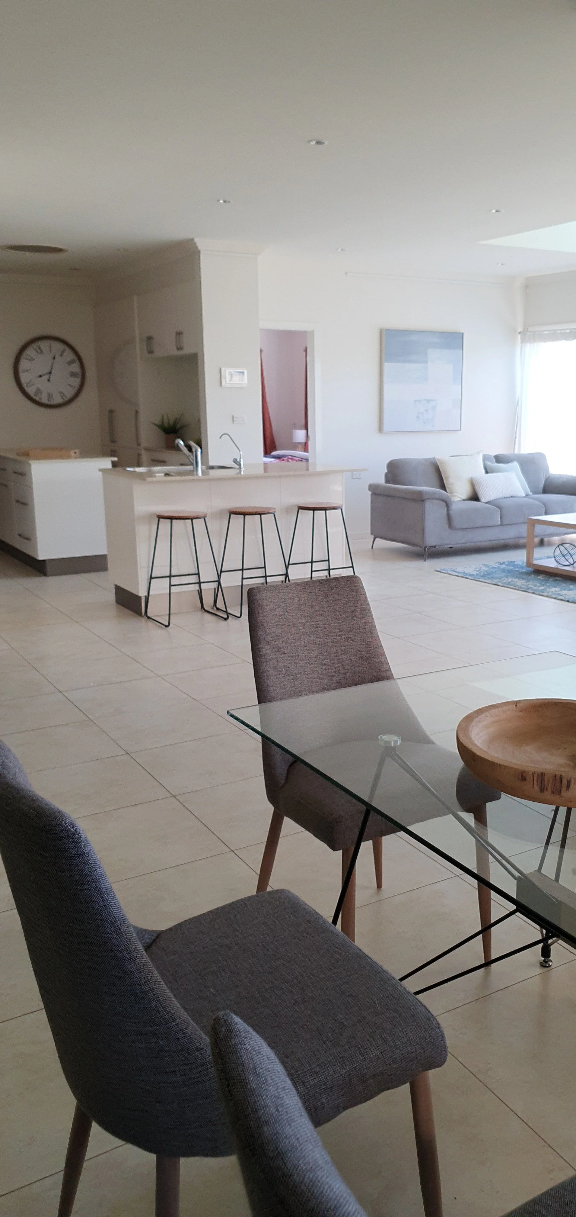 Modern open-concept living room and kitchen with tile flooring, gray sofa, dining table with chairs, and kitchen island with bar stools.