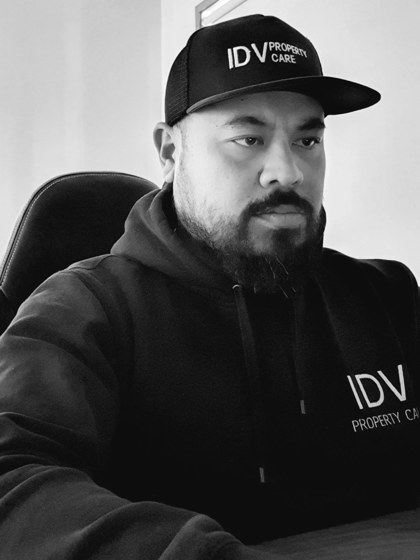 A man with a beard wearing a black hoodie and a black cap with white text that reads 'IDV Property Care' sitting in a black office chair against a plain wall.