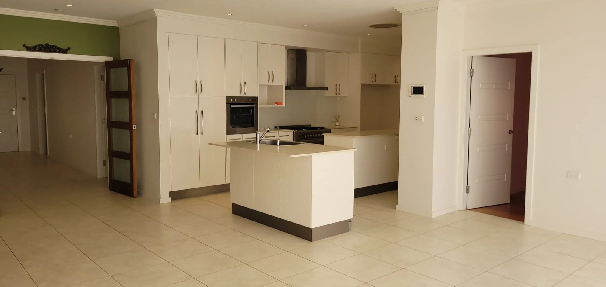 Empty modern kitchen with white cabinets, a small island, oven, stove, and stainless steel sink, open floor plan with beige tiled floors.