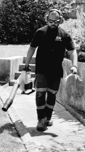 A man walking on a sidewalk, wearing safety glasses, headphones, and work uniform with reflective strips, holding a leaf blower.