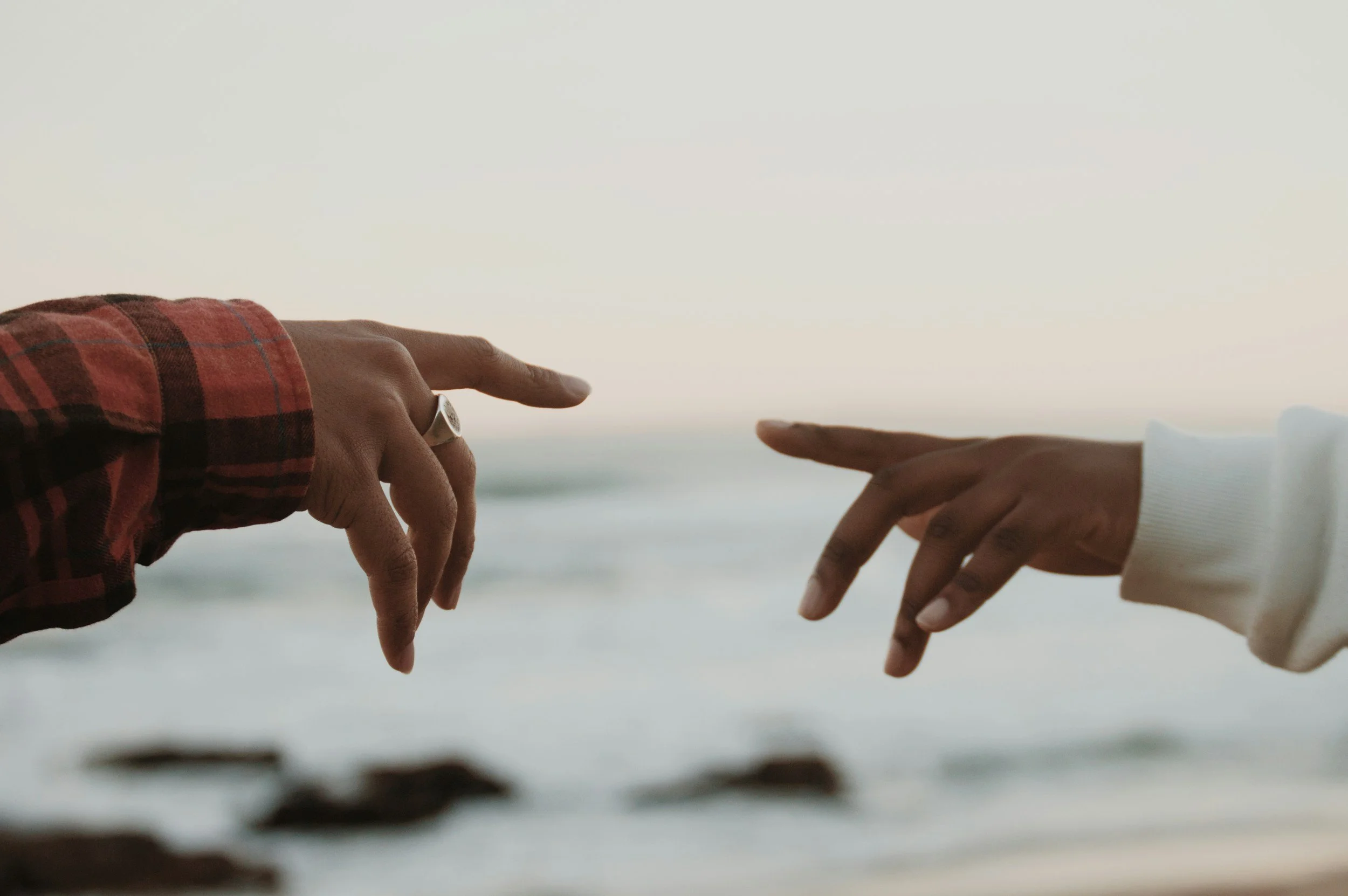 Two hands reaching towards each other, one wearing a red plaid sleeve and the other a white sleeve, against a calm ocean background during sunset.