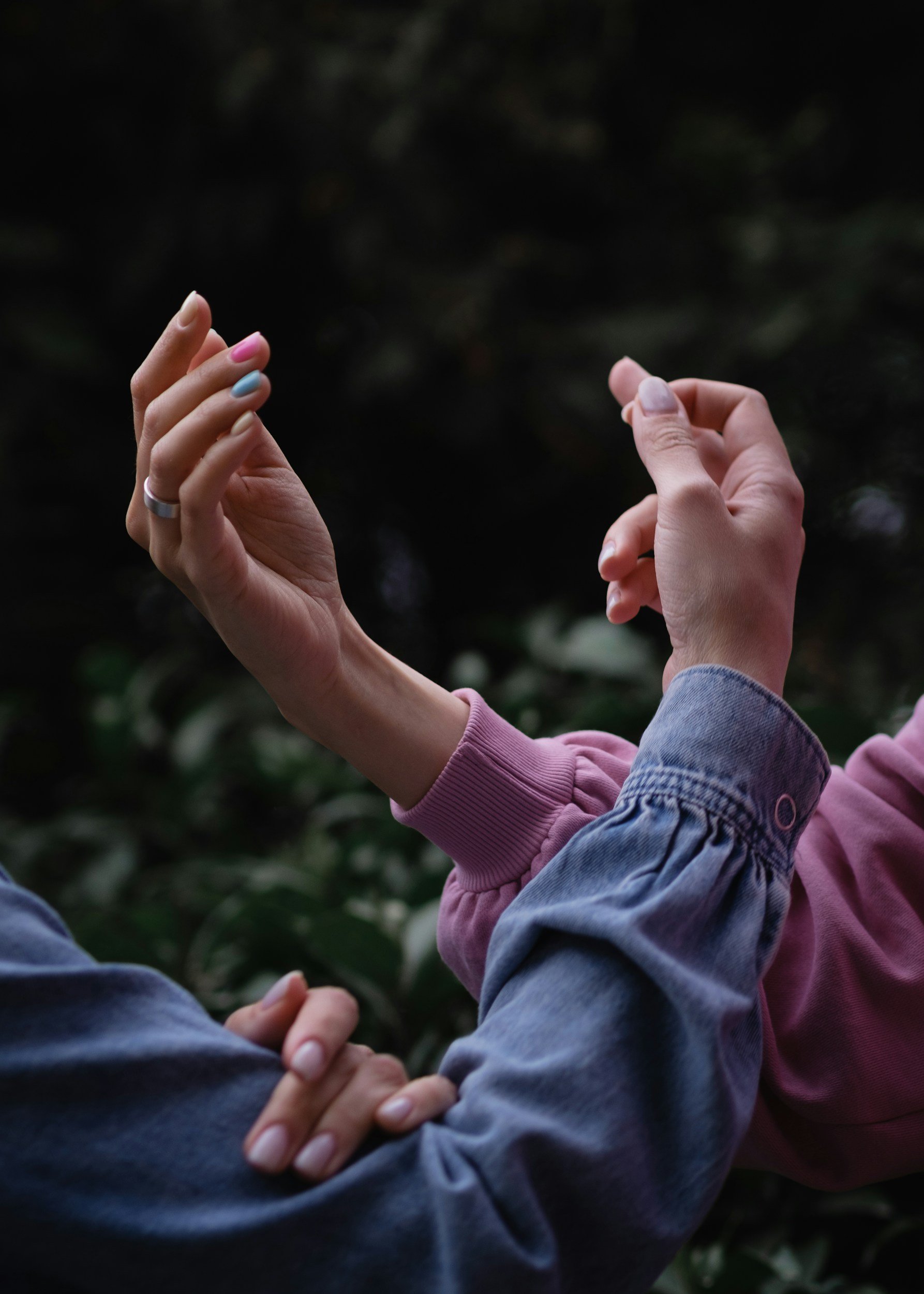 Close-up of two hands, one with colorful nail polish and a ring, reaching toward another hand with natural nails. They appear to be holding or about to hold each other against a dark, blurred background.