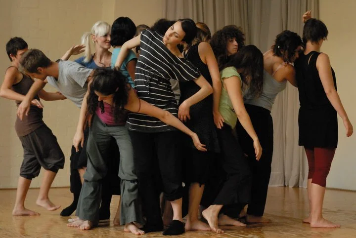 A group of dancers performing a contemporary dance piece on stage, with some dancers leaning on each other and others standing apart, all dressed in casual dance attire.