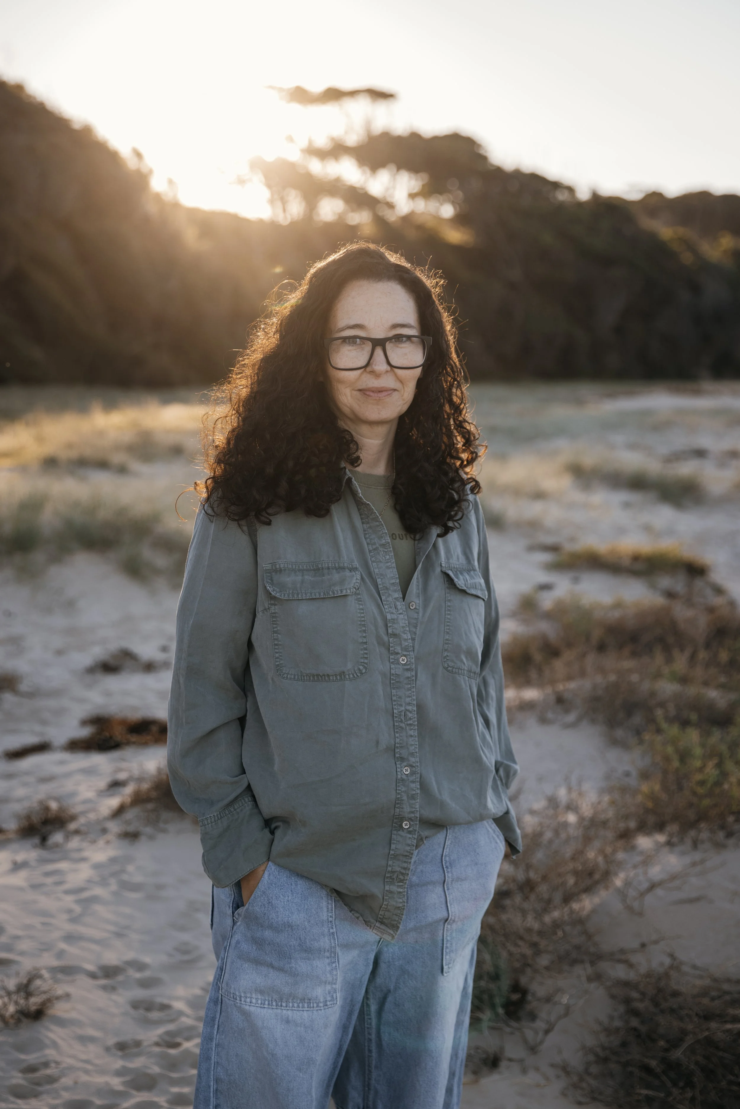 A woman with curly dark hair, glasses, wearing a gray jacket and blue jeans, standing outdoors on a sandy area with bushes and trees in the background, during sunset.