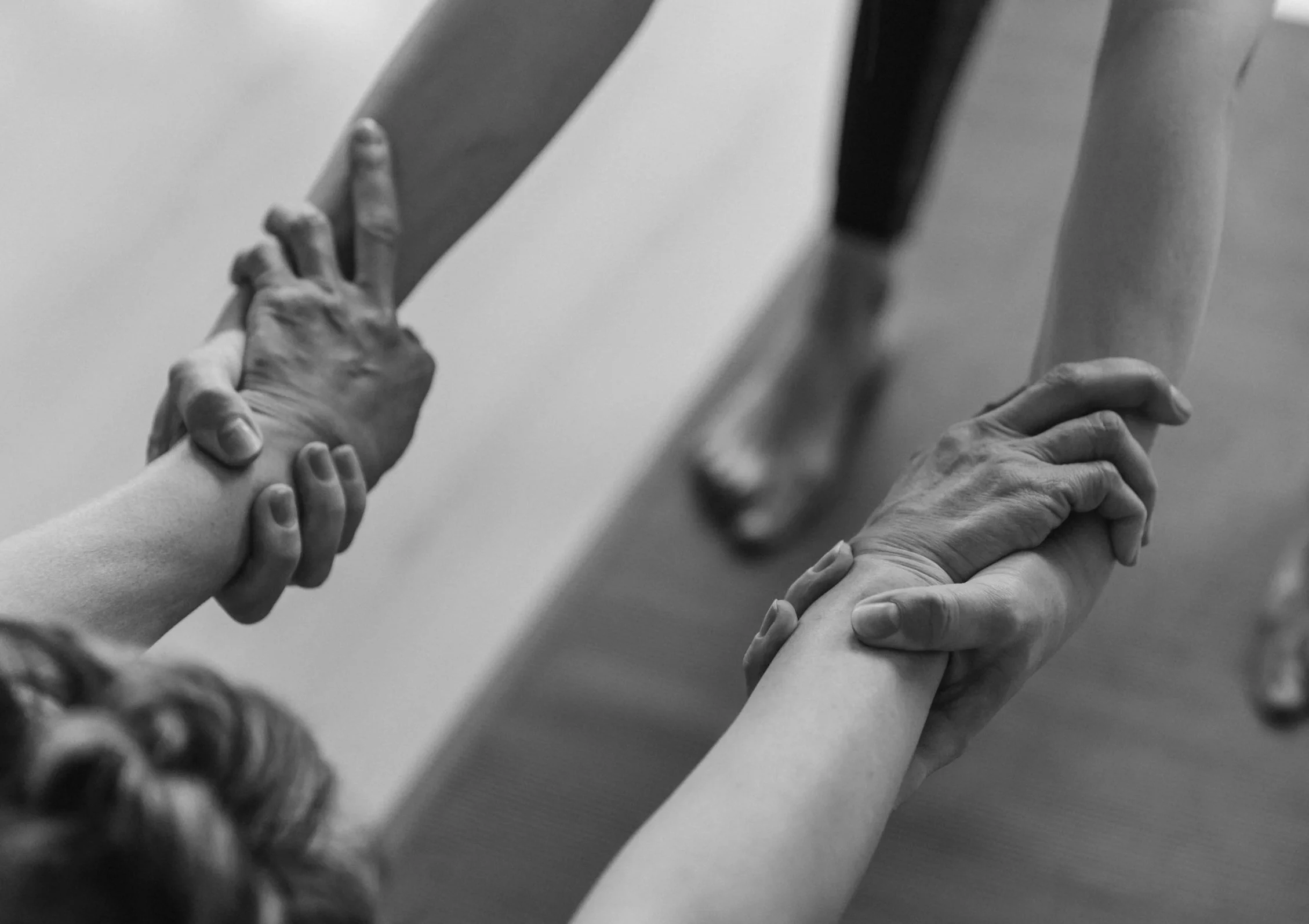 Black and white photo of multiple hands, some with bracelets, holding and supporting each other's arms and wrists, creating a interconnected and caring gesture.