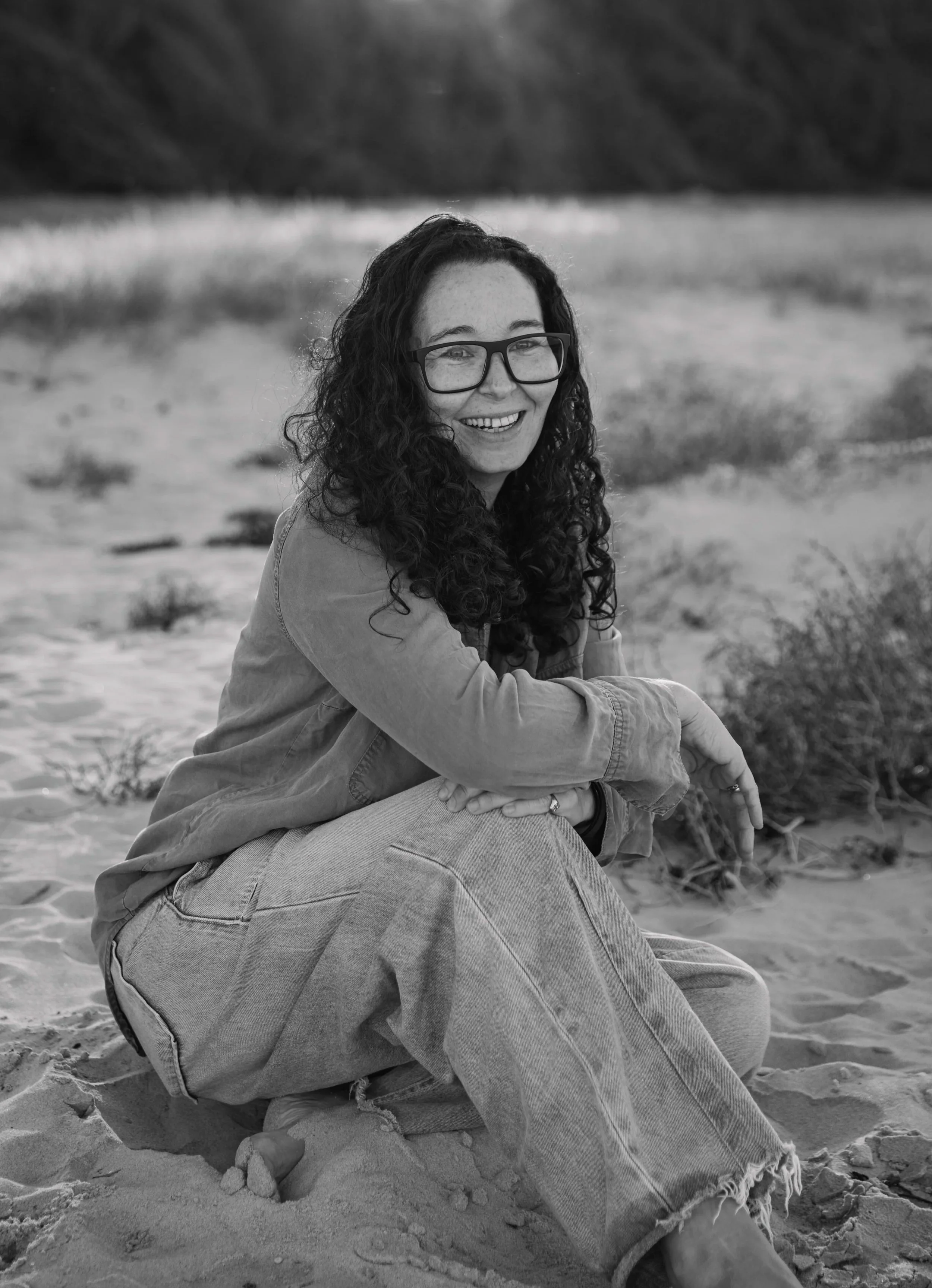 A woman with curly hair and glasses sitting on the sand at the beach, smiling, with a blurred shoreline and trees in the background, in black and white.