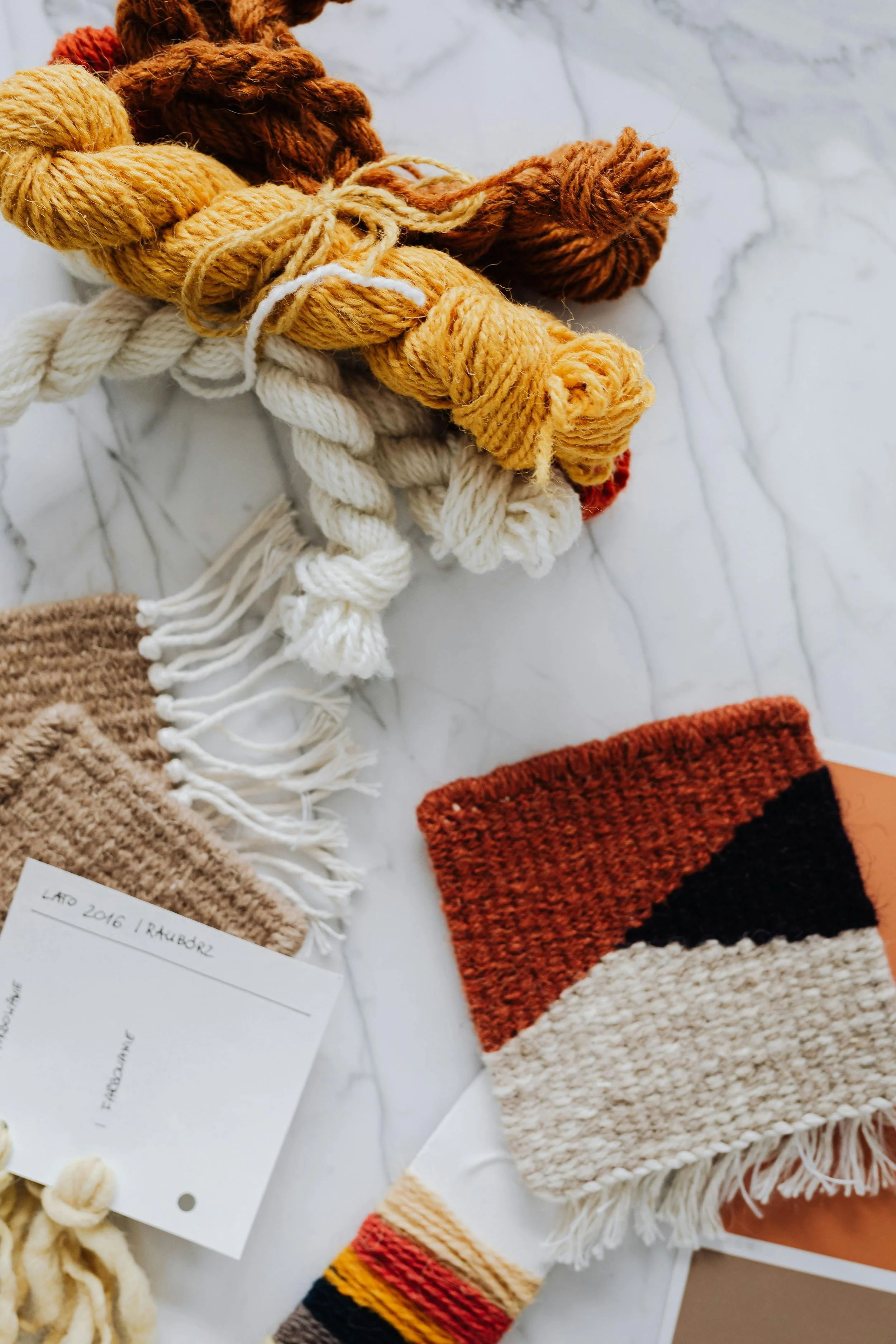 Various woven and knitted textile items including a beige fringed scarf, a white fringed scarf, a multicolored striped sock, and a warm hat, displayed on a white marble surface.