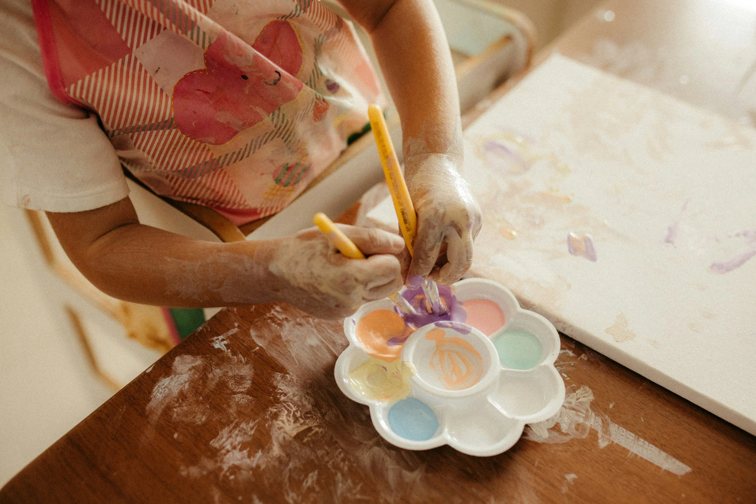 A child wearing a pink apron with a butterfly design is painting using watercolors on a wooden table. Several small watercolor paint palettes are visible, with various pastel colors. The table has some paint splatters and smudges.