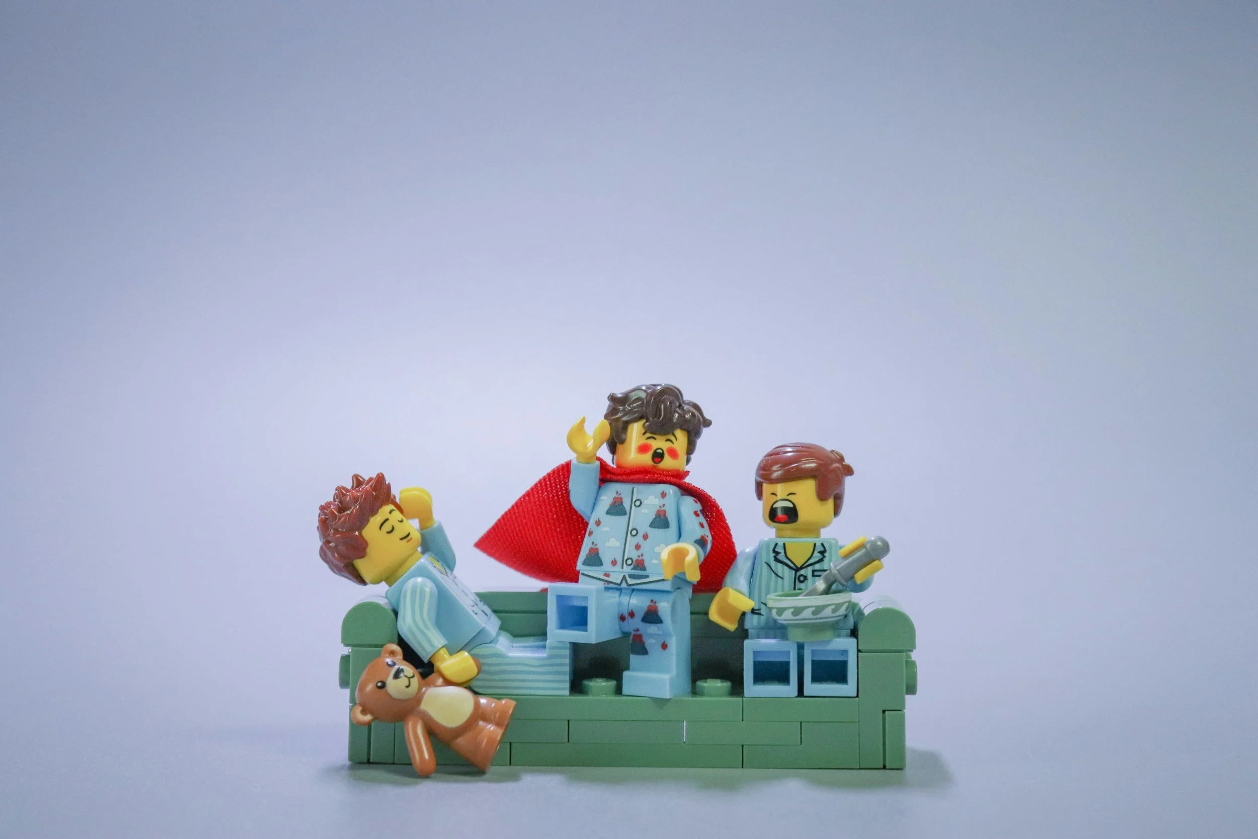 LEGO figurines depicting a family scene, with a woman in pajamas and a red cape sitting on a green couch, surrounded by children and a lion plush toy, against a plain background.