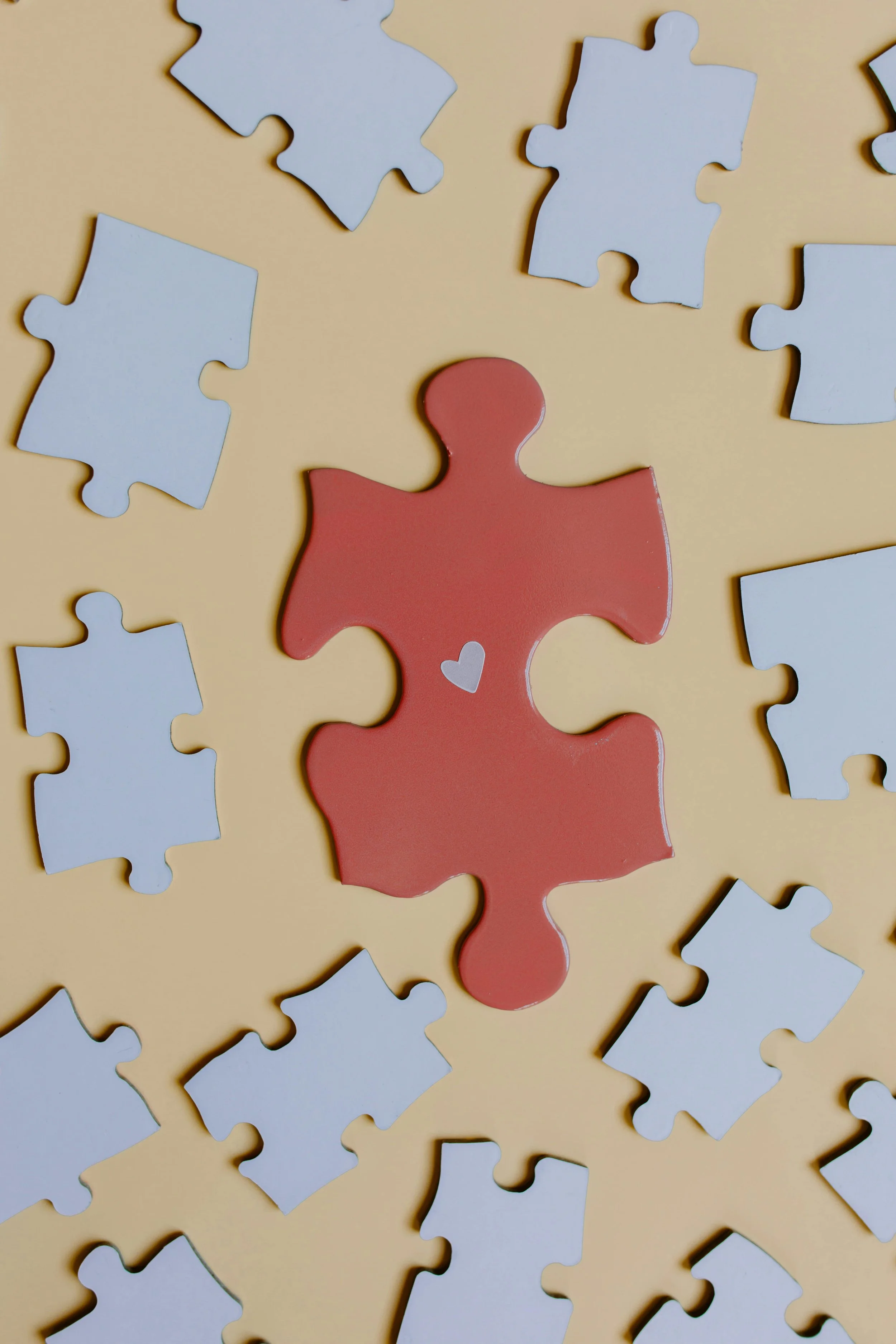 A red puzzle piece with a small white heart on it, surrounded by white puzzle pieces on a beige background.