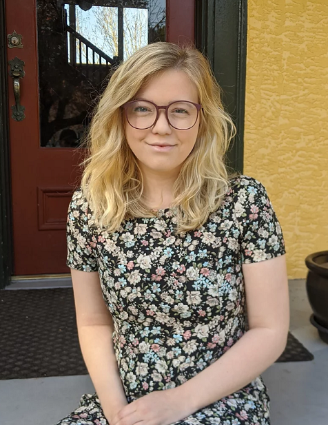 A young woman with blonde, wavy hair and glasses sitting outside in front of a yellow wall and dark red door, wearing a floral dress.