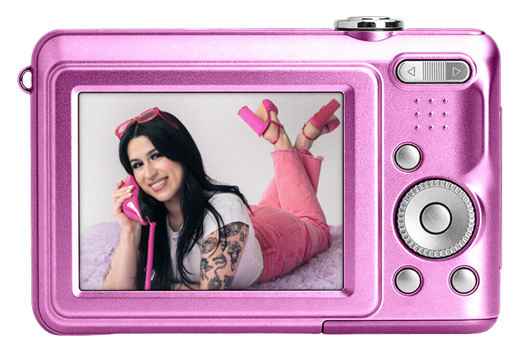 A pink digital camera displaying Gabi, founder of The Estie Edit, with dark hair, tattoos, smiling and talking on a pink rotary phone, lying on a soft surface in a playful pose with pink high-heeled shoes.
