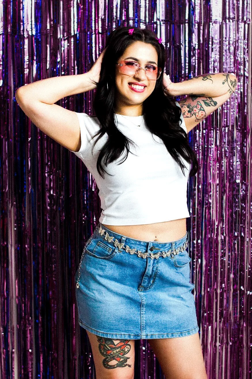 Gabi, founder of the Estie Edit, is smiling with glasses and tattoos, wearing a white crop top and denim skirt, standing in front of purple metallic curtain.