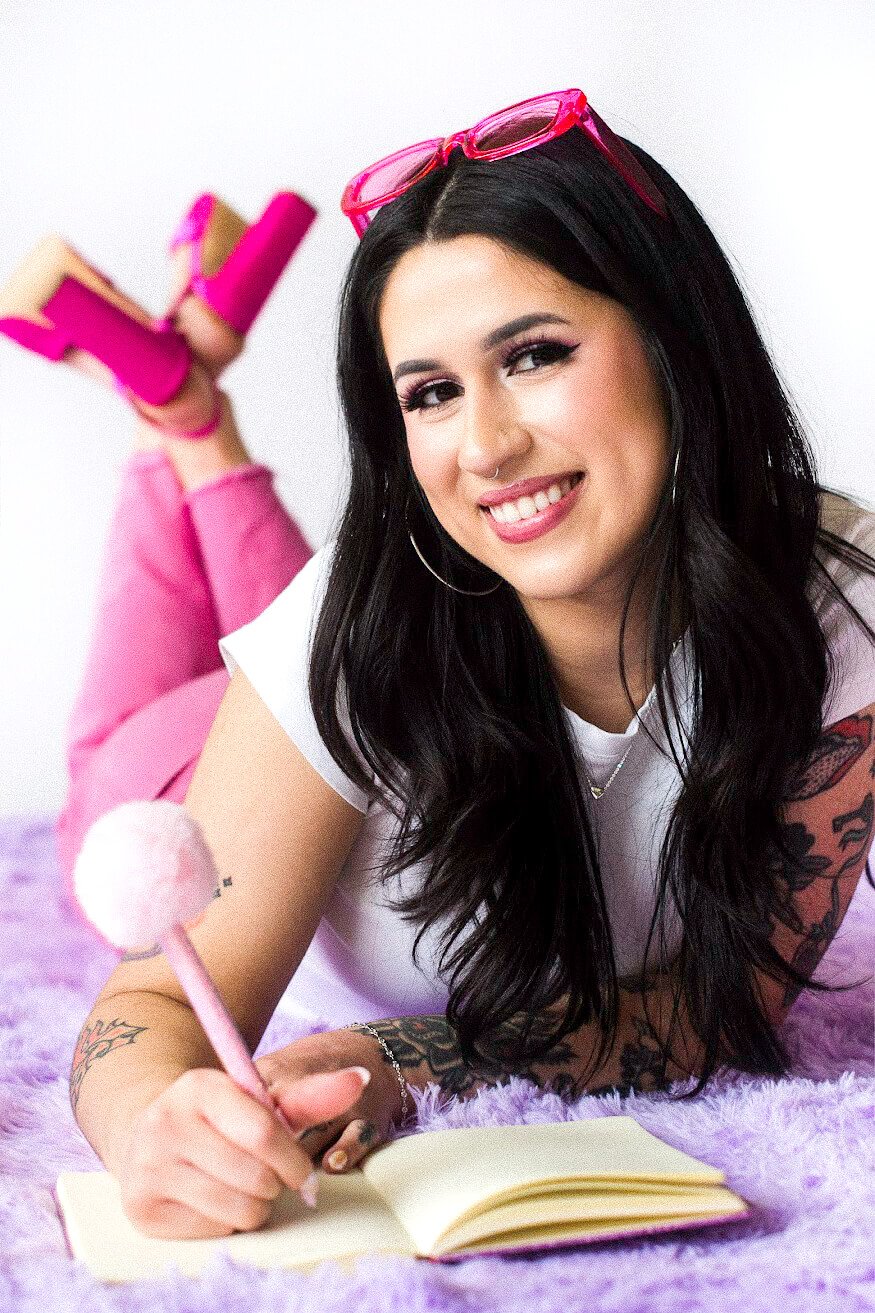 Gabi, founder of The Estie Edit, has long black hair, tattoos, and pink sunglasses on her head. She is lying on a purple fuzzy surface, smiling at the camera while writing in a small notebook with a pink pen.