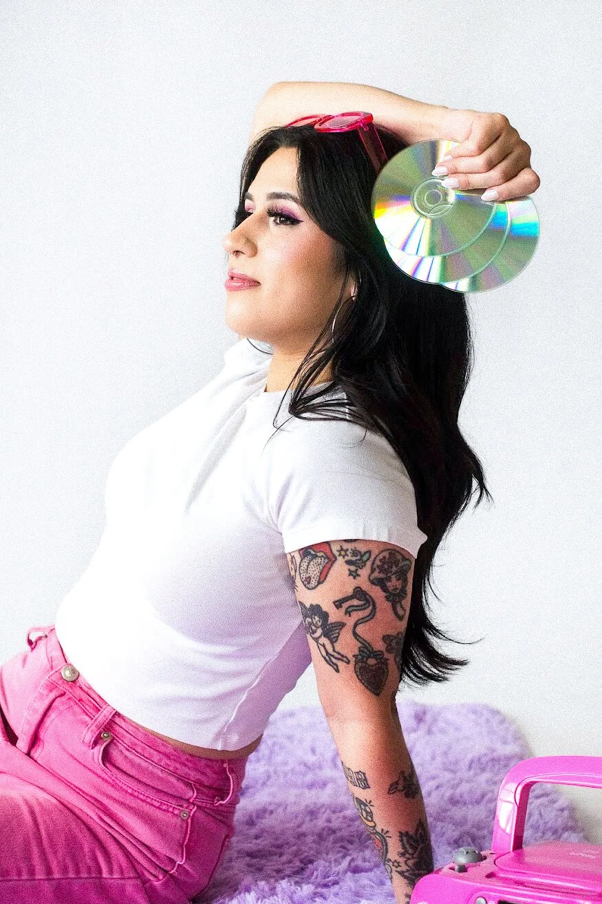 Gabi, found of The Estie Edit, has long dark hair, tattoos on her arm, and is wearing hot pink pants. She is holding two compact discs near her head, against a plain white background.