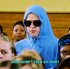 Scene from the movie Mean Girls shows a person wearing sunglasses and a blue hoodie with the hood up, surrounded by other students, with a subtitle that reads 'SHE DOESN'T EVEN GO HERE!'