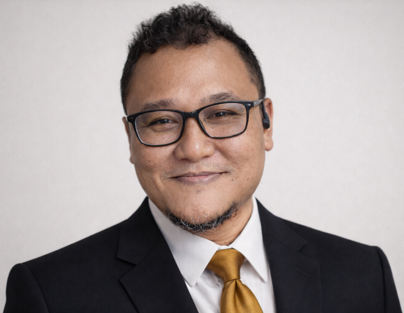 Headshot of an Asian man wearing glasses, a black suit, white shirt, and gold tie, smiling against a plain background.