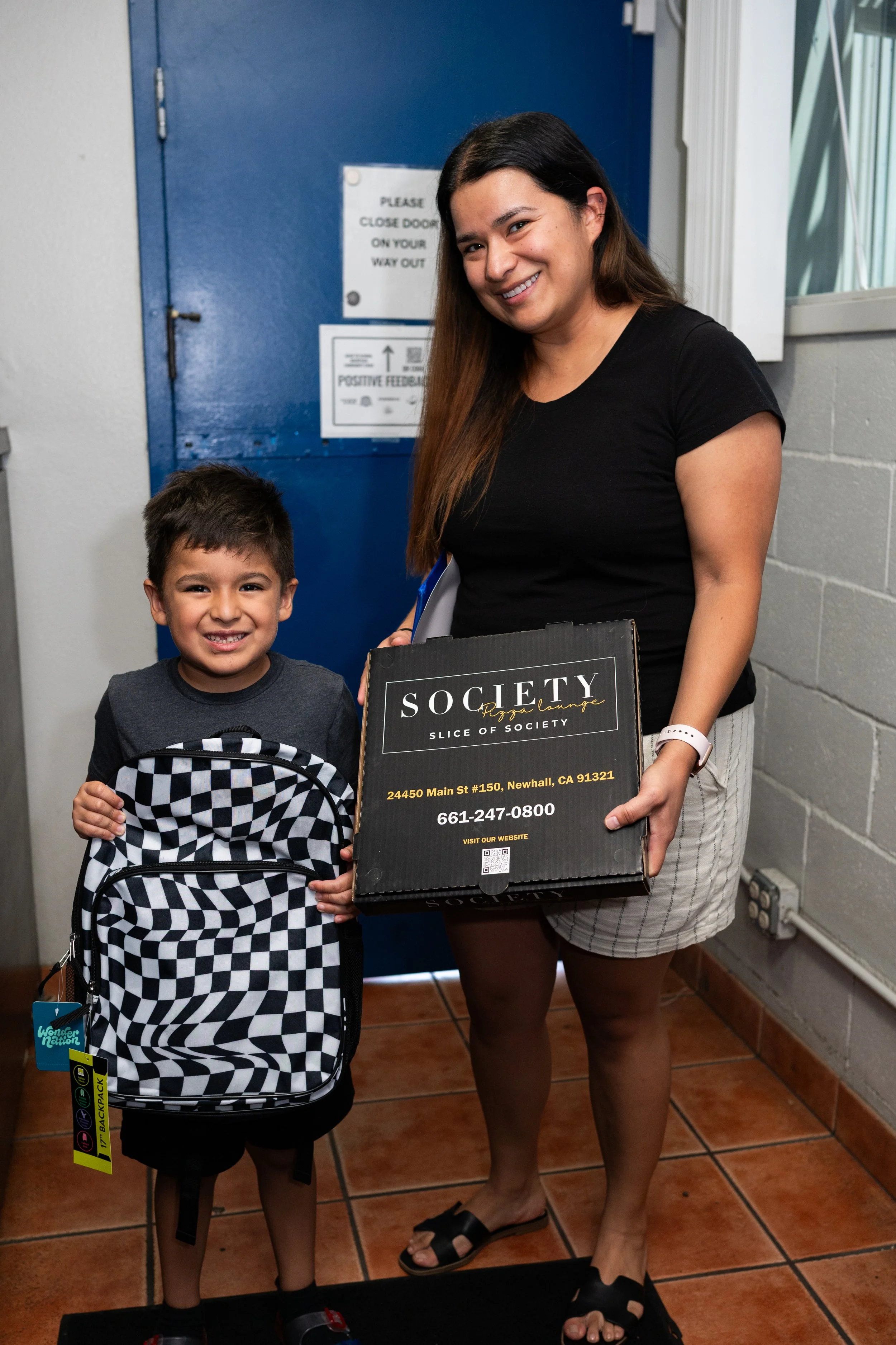 A woman and a young boy standing indoors, smiling at the camera. The boy holds a black and white checkered backpack. The woman holds a black box with white text advertising a pizza lounge and an address in Newhall, California.