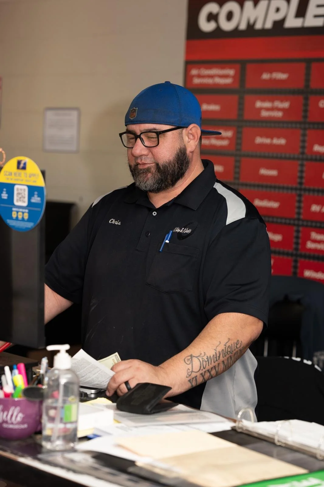 A man wearing a blue cap, glasses, and a black uniform with gray accents stands behind a counter, handling money and a receipt, with various office supplies and sanitizer on the counter. A red and black menu board is visible in the background.