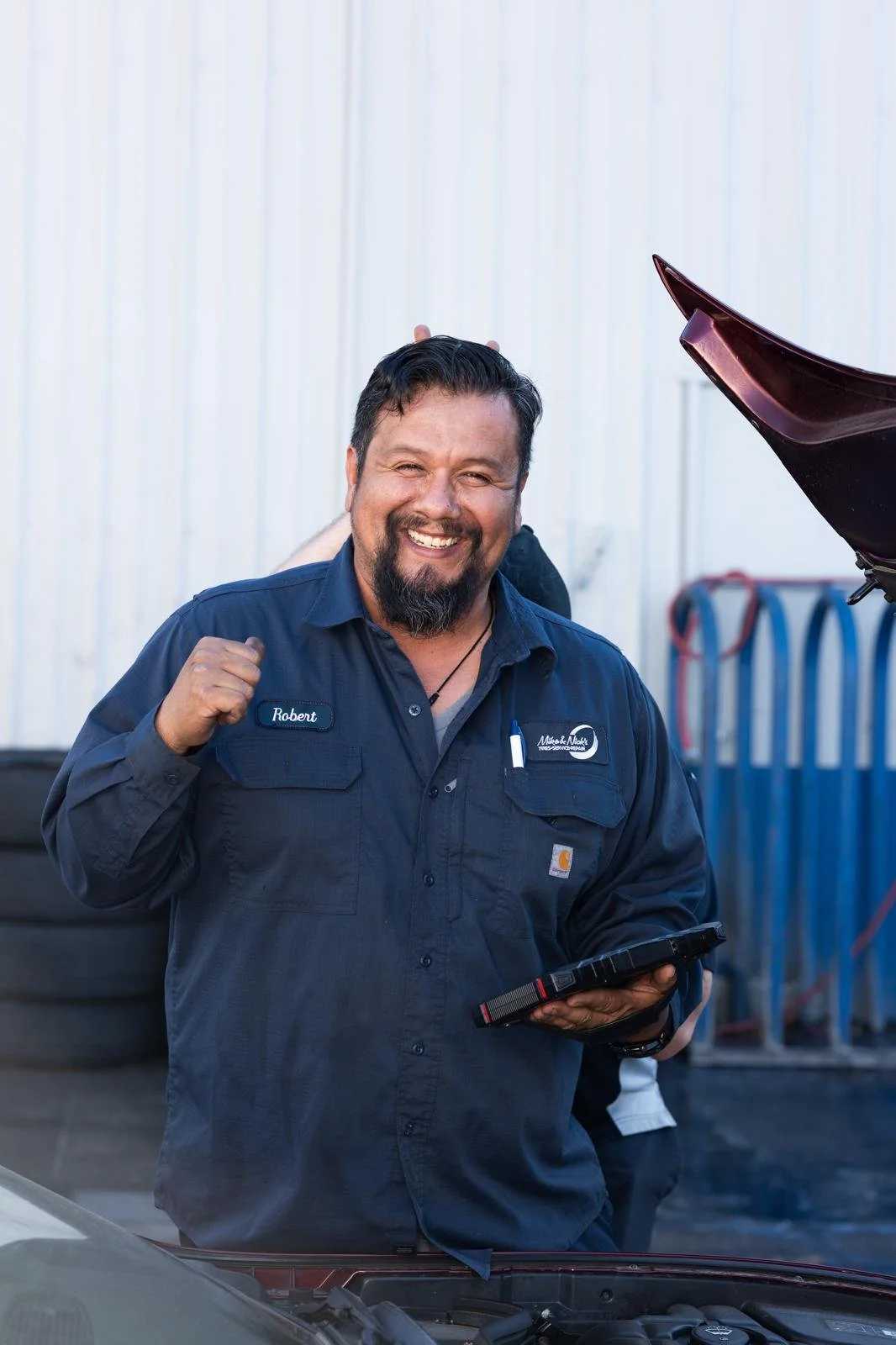 A happy man with a beard and dark hair, wearing a blue mechanic uniform with the name tag Robert, giving a fist pump in front of a car repair shop.