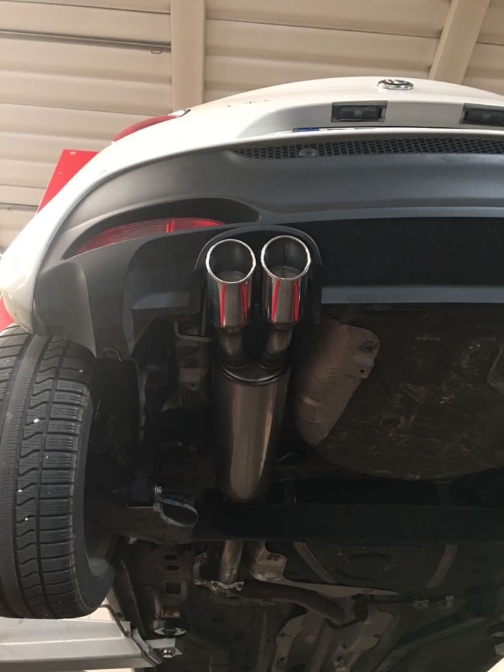 The rear underside of a car shows a dual exhaust pipe with shiny chrome tips, tire, and part of the undercarriage.
