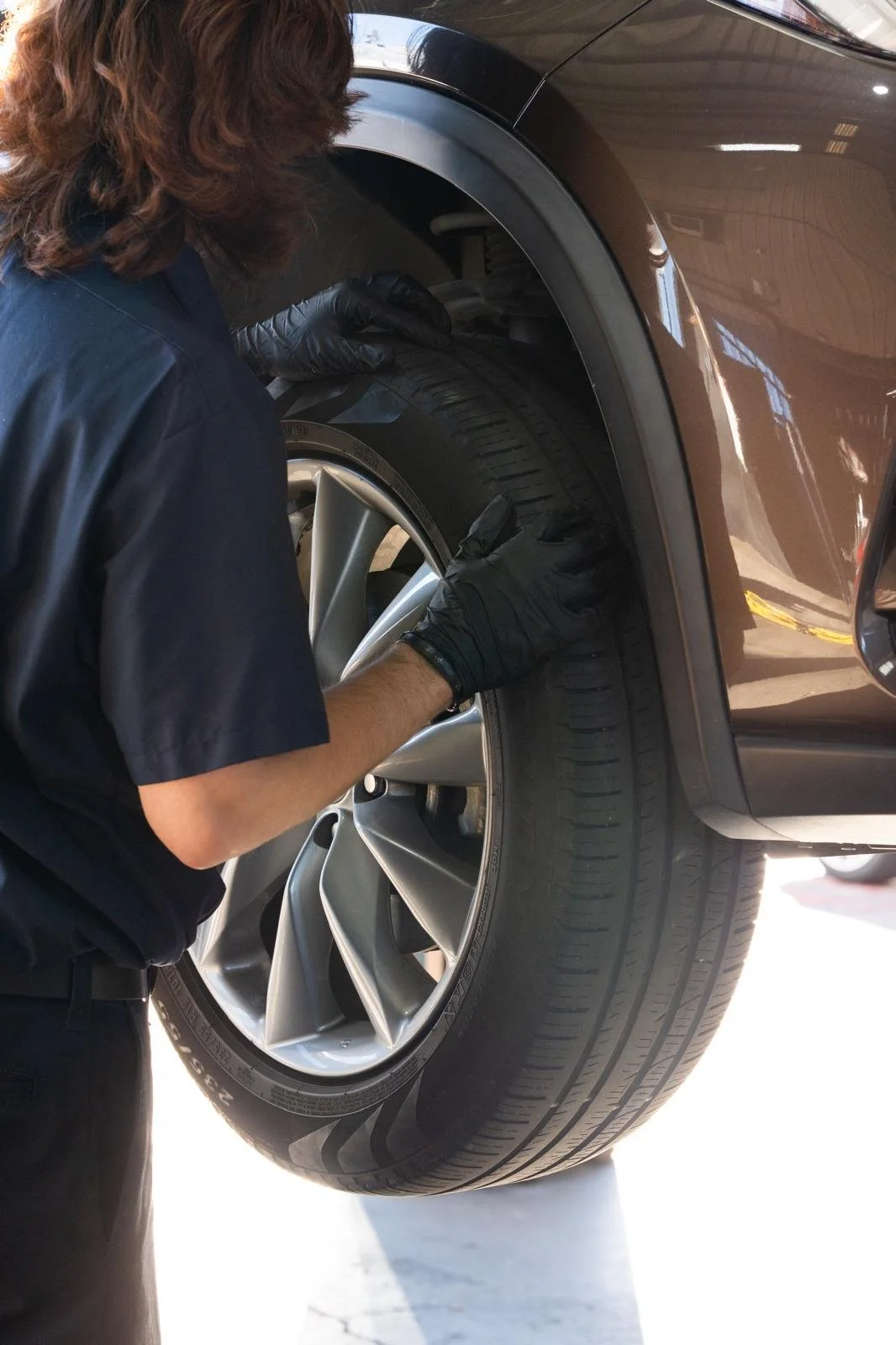 A person in black gloves inspecting the front tire of a brown car in a garage.
