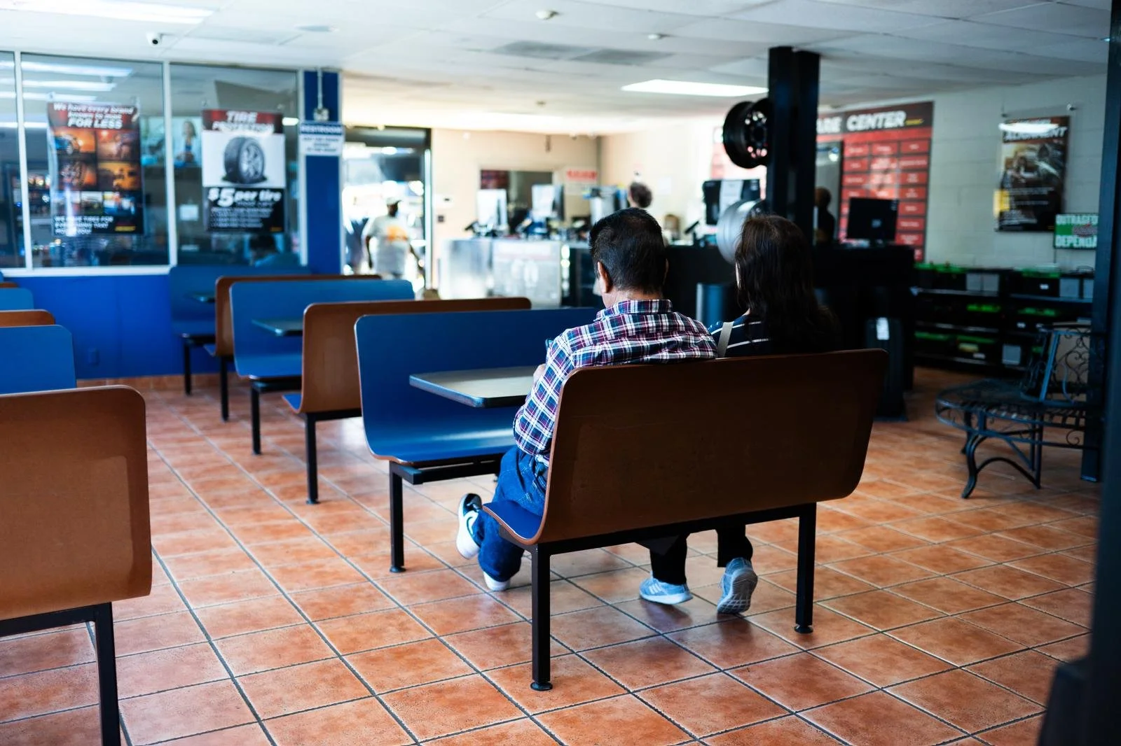 Interior of a fast-food restaurant with empty tables and a couple sitting on a bench, facing away from the camera, with a counter in the background.