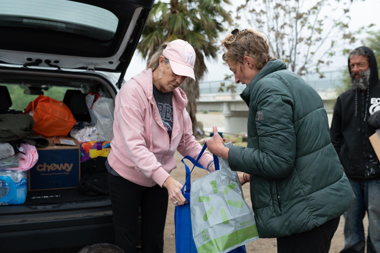 Two women exchanging a plastic shopping bag outdoors next to an open vehicle trunk filled with supplies, with a man in the background observing, under a cloudy sky.