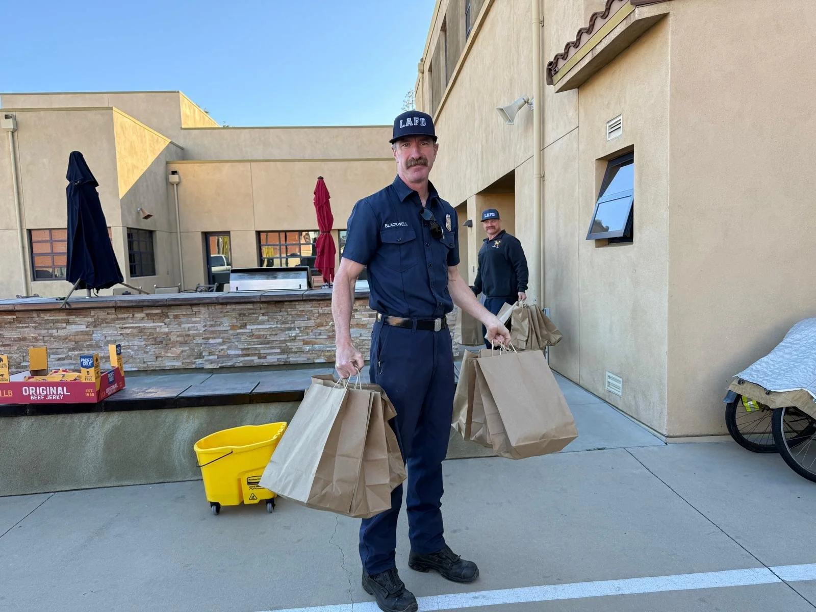 A police officer with a mustache wearing a dark blue uniform and a cap labeled 'LAFD' is holding multiple brown paper bags, standing outdoors in front of a beige building with a brick counter and a yellow mop bucket nearby. Another officer is visible