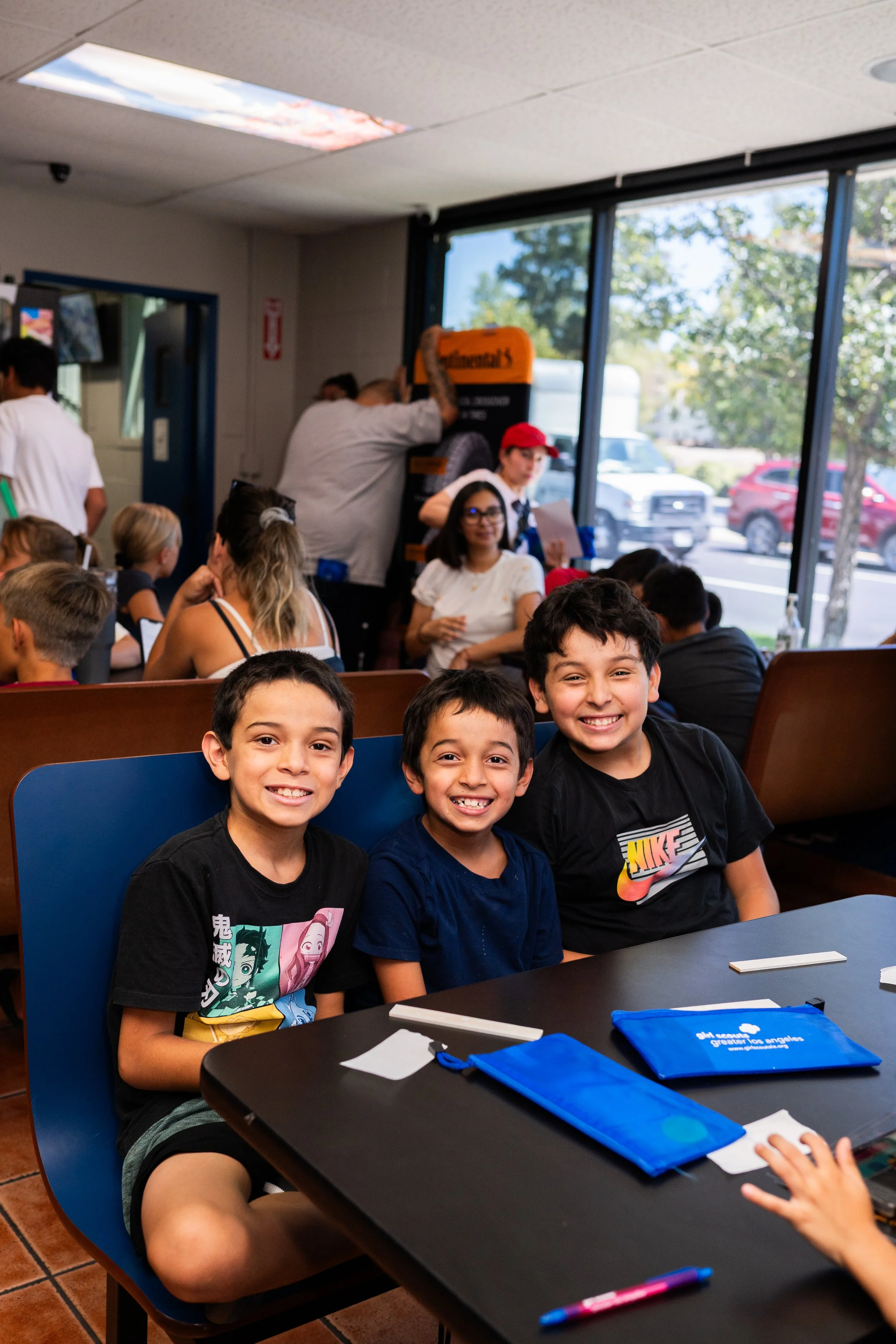 Group of kids sitting at tables in a restaurant or cafe, smiling at the camera, with some adults and a staff member in the background near a window.